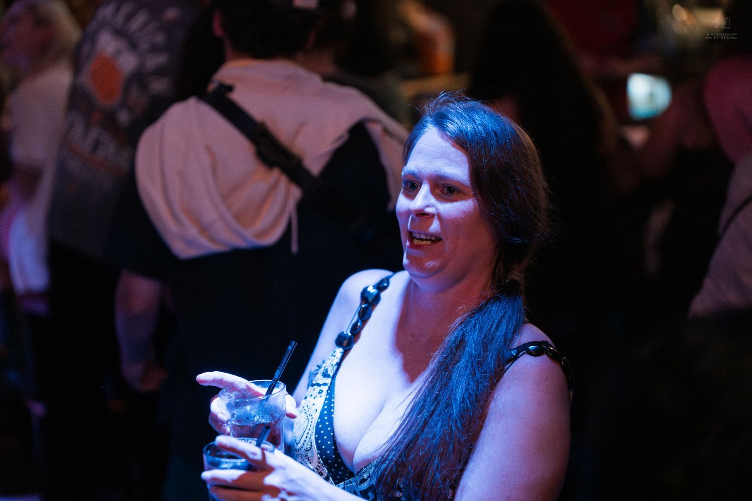 Woman with dark hair smiles while holding a cocktail glass at a dimly lit bar or nightclub.
