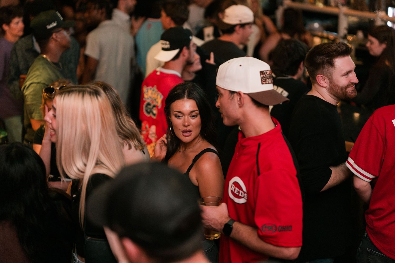 Young adults socializing in a packed bar, with a woman in a black top talking to a man in a red Reds jersey.