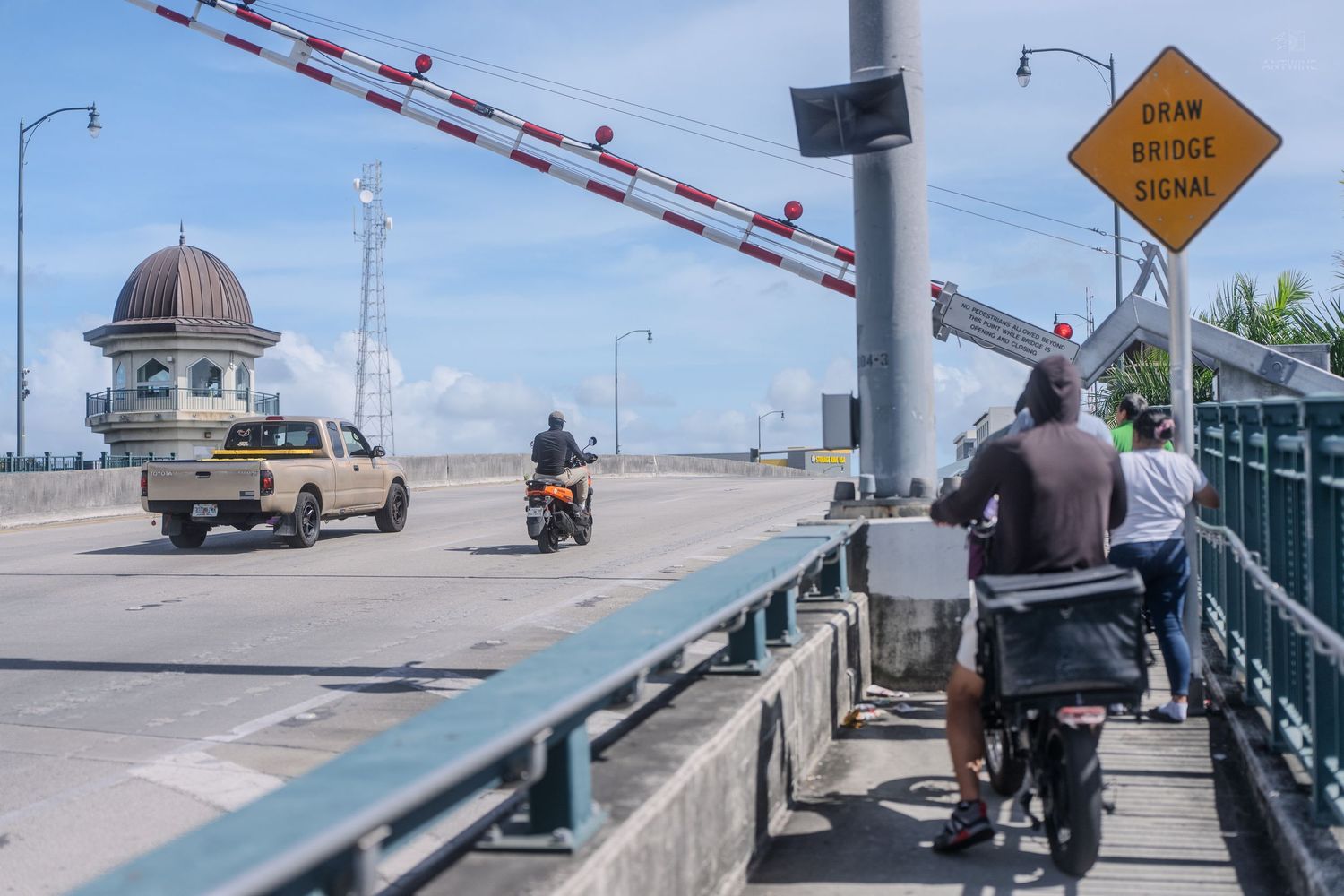 Motorcyclists and truck crossing a drawbridge with lowered gate arm and draw bridge closed sign.