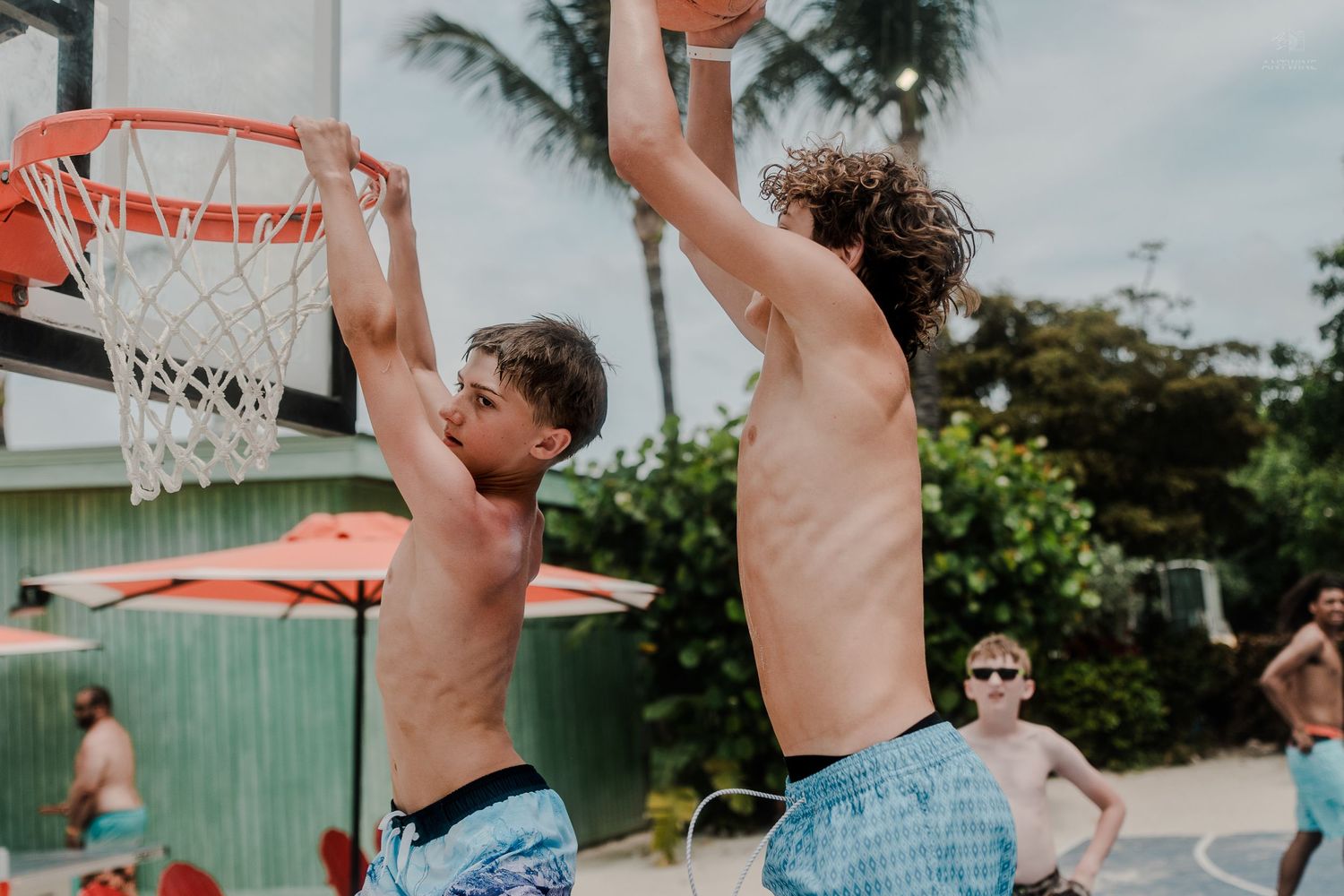 Two shirtless boys playing basketball outdoors at a tropical resort court.