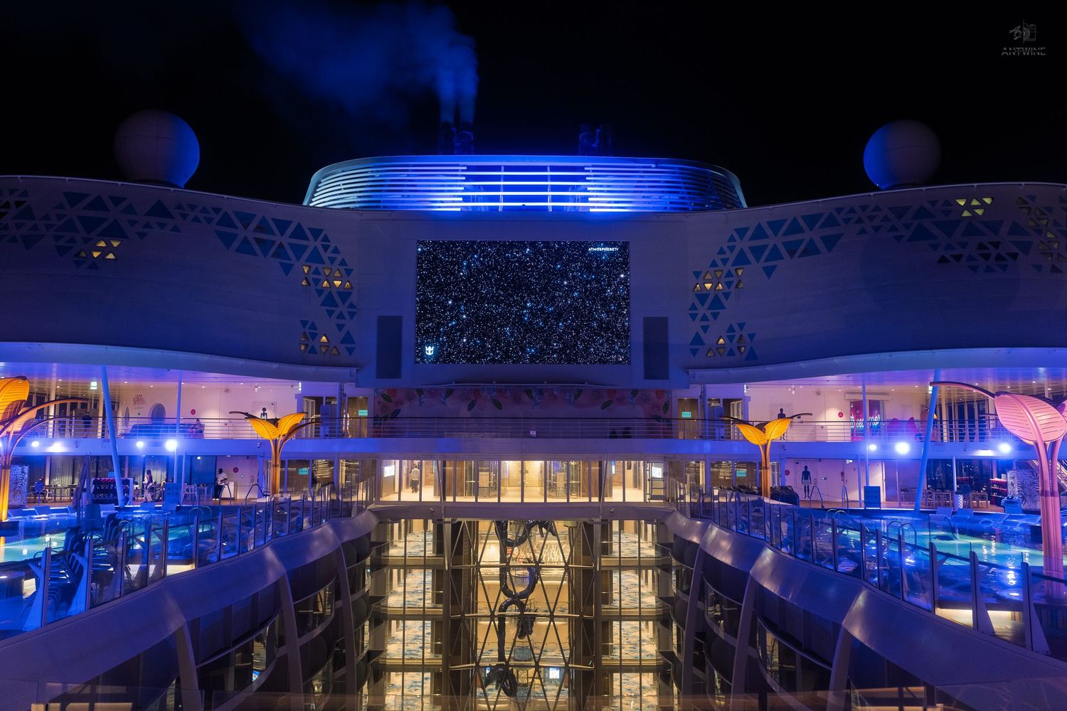 Cruise ship deck illuminated with blue lights at night featuring a large screen and pool area.