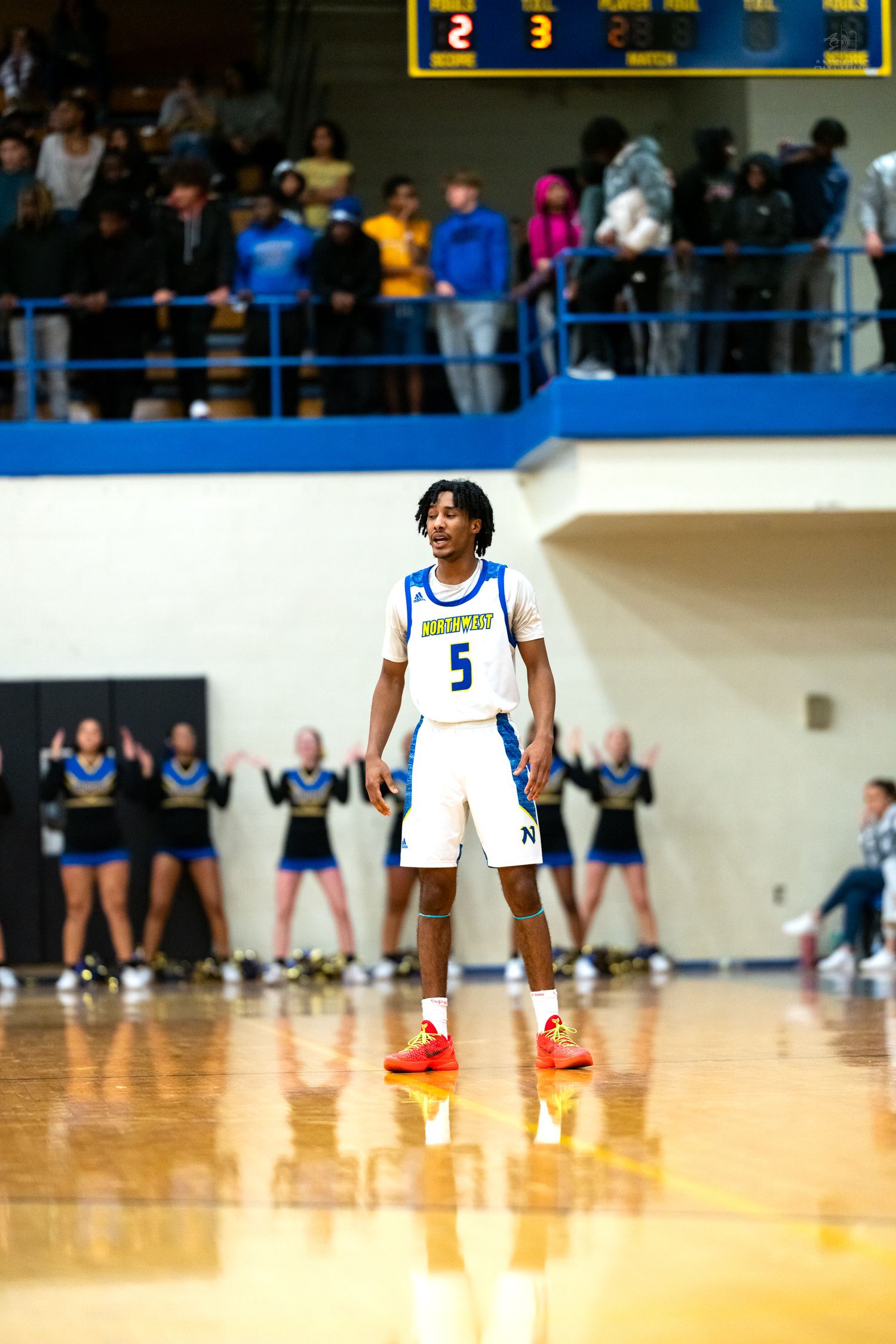 Basketball player #5 in white uniform stands at free throw line with cheerleaders behind.