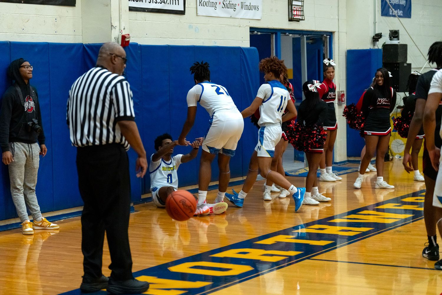 A basketball player sits on the court near the baseline as teammates and cheerleaders look on during a game.