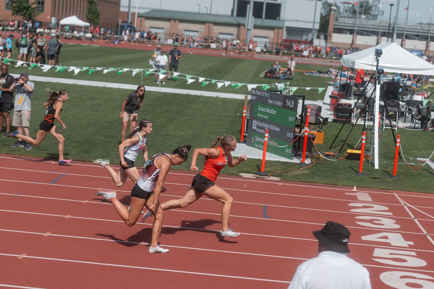 Female track athletes race toward finish line on red track at sunny outdoor high school championship meet.