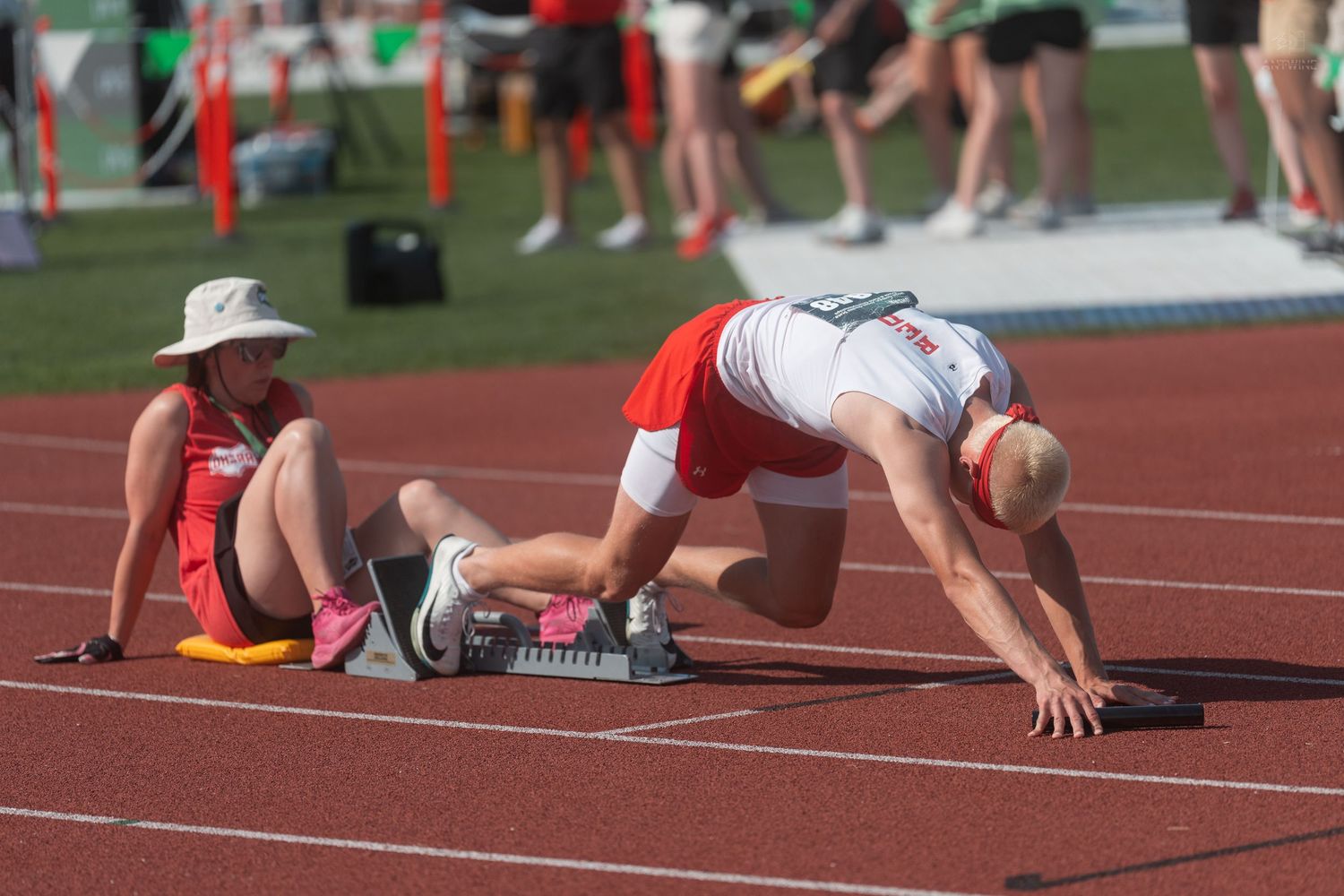 Track athlete in red uniform crouches in starting position on blocks while official sits nearby on track.