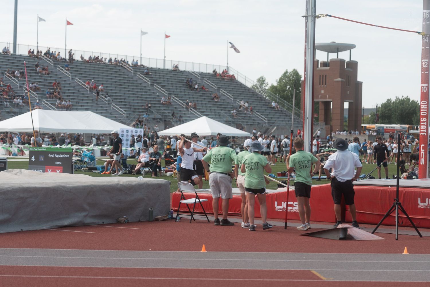 Officials in green shirts manage a field event at a packed track and field stadium on a sunny day.