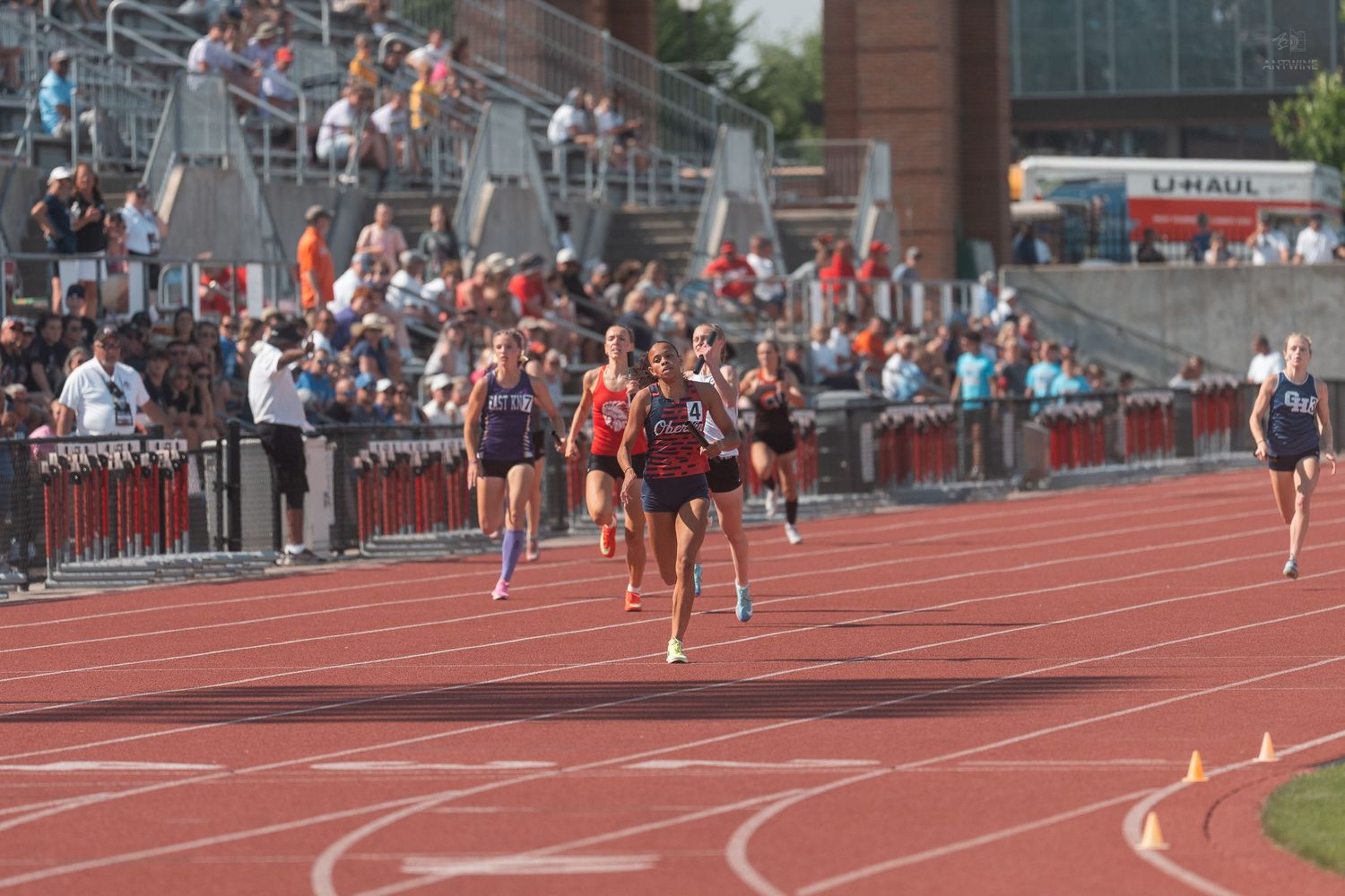 Female athletes compete in a distance running race on a red track with large crowd watching.