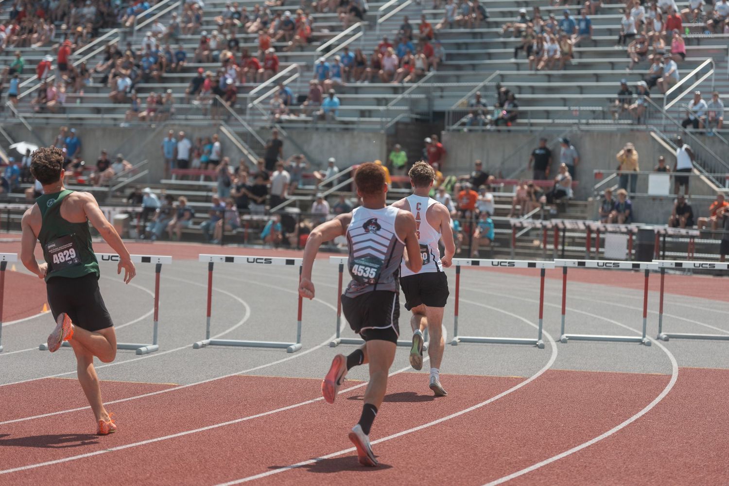 High school athletes sprint toward hurdles on a red track with a packed grandstand visible in the background.