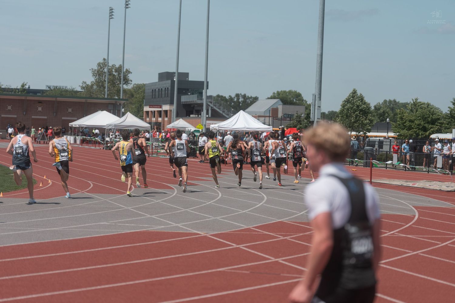 Athletes race on a red track at an outdoor track and field competition with white tents in background.