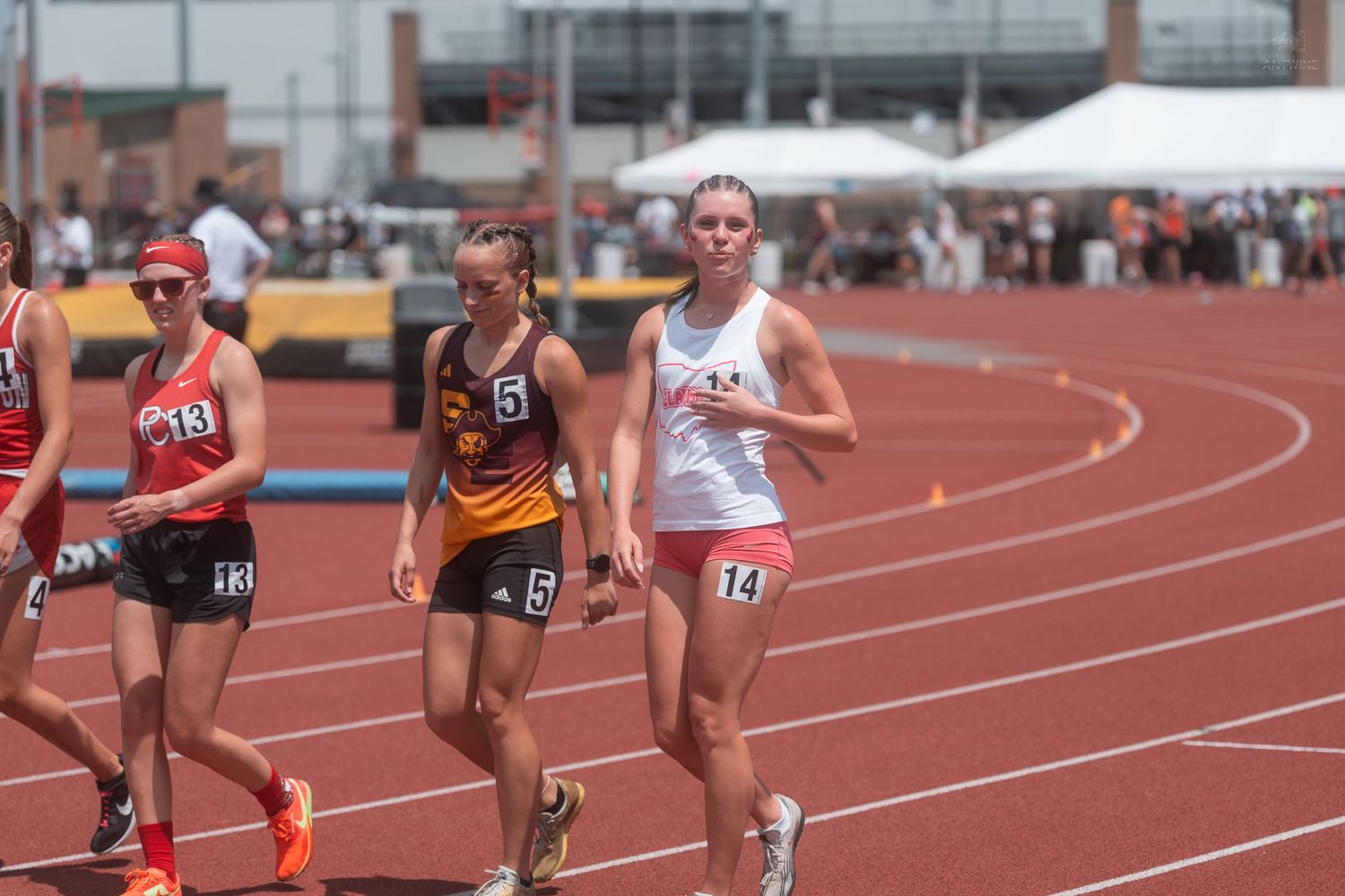 Female track athletes wearing numbered bibs racing on red outdoor track during sunny high school competition.