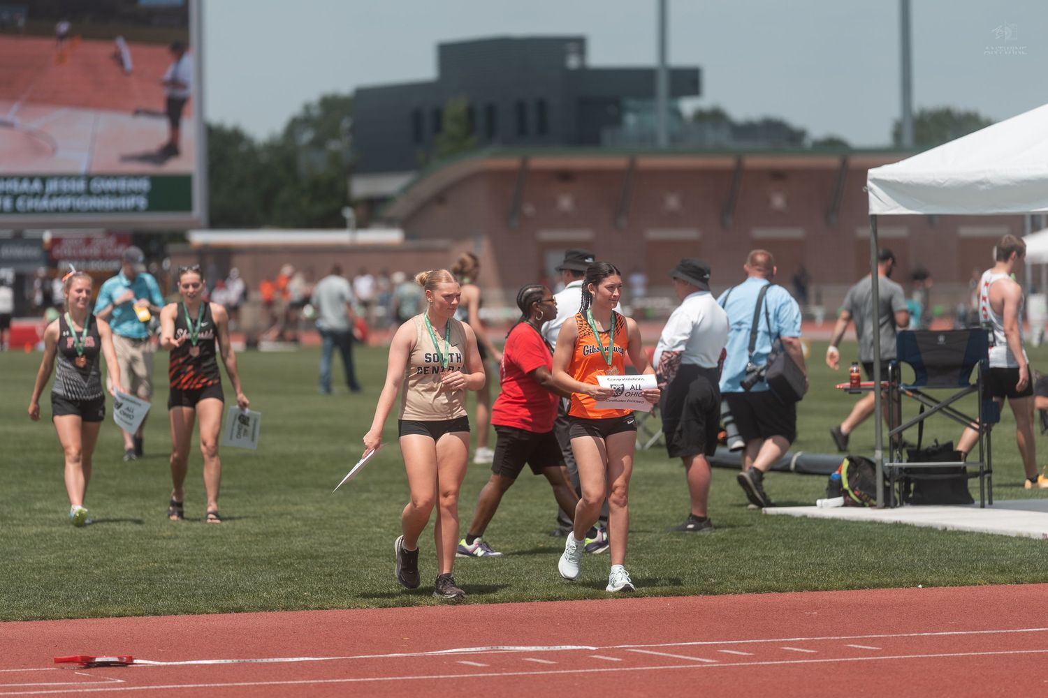 Female track athletes walk along the infield track area at the OHSAA Track and Field State Championships on a sunny day.
