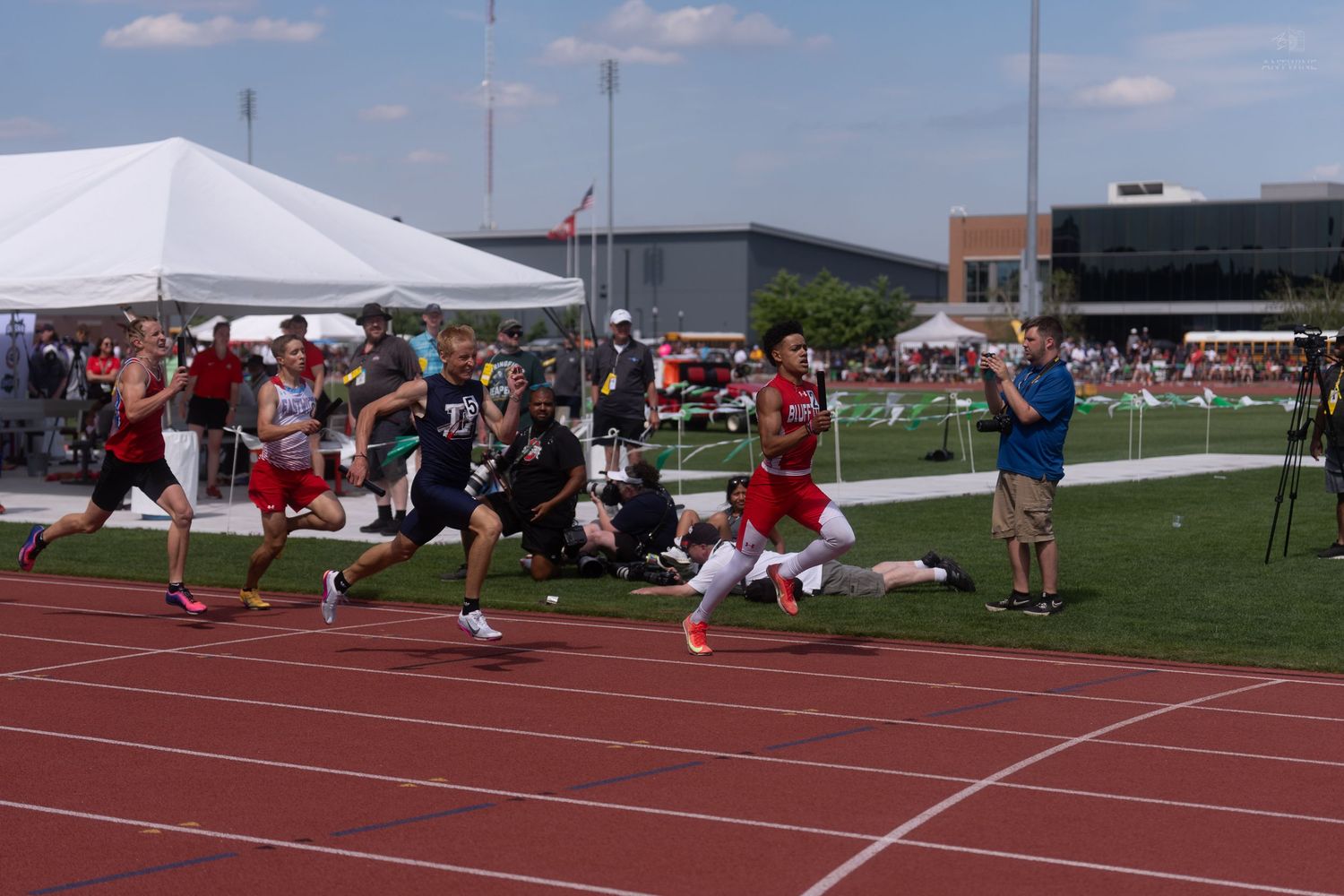 Group of athletes race toward finish line at outdoor track meet with spectators and photographers on sidelines.