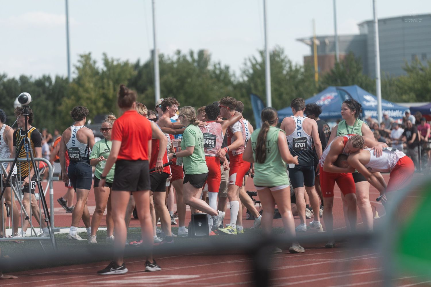 Groups of track and field athletes in various uniforms standing together on a red running track.
