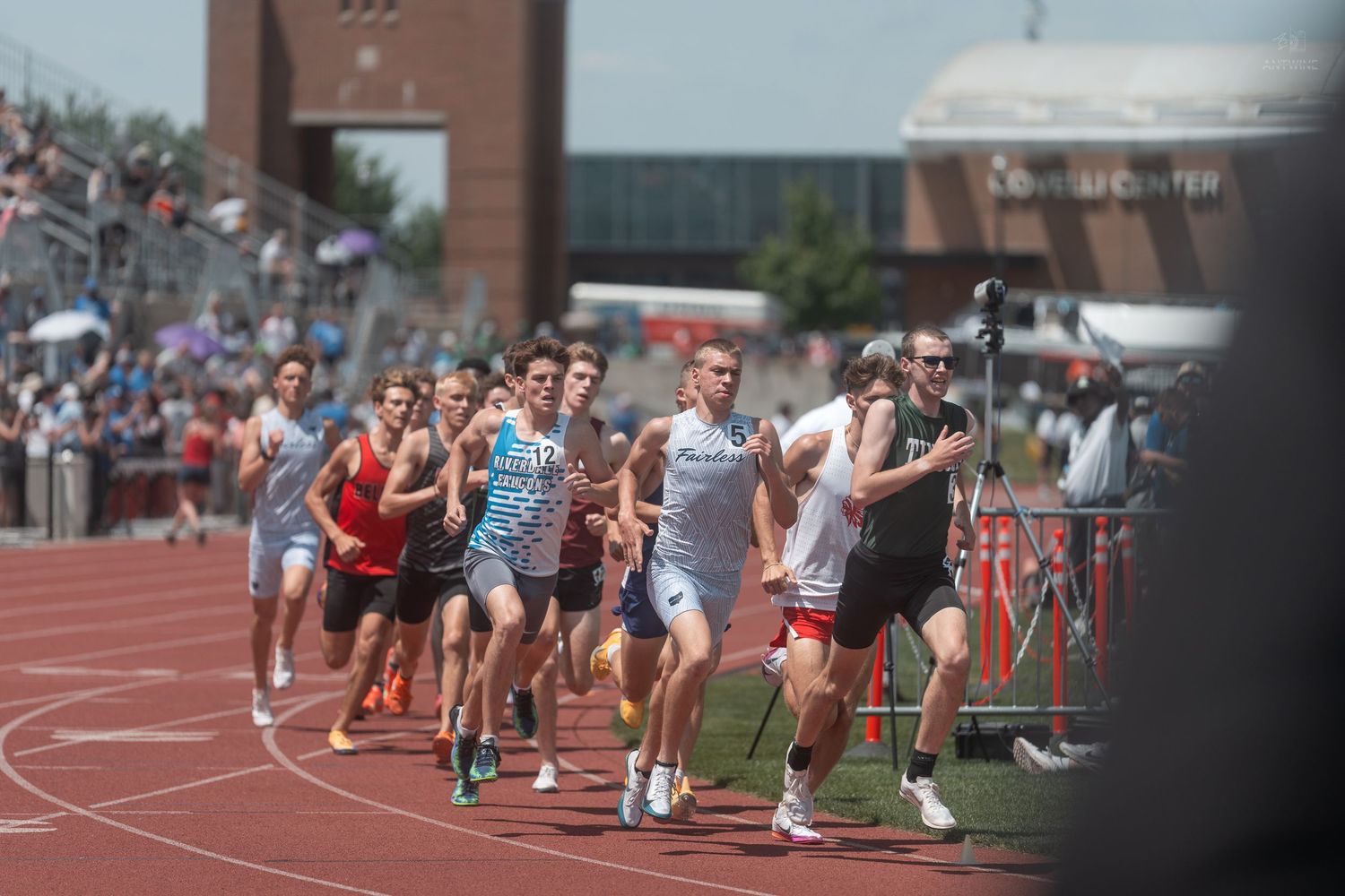 Male runners compete in a track race at an outdoor stadium during a sunny day athletic meet.