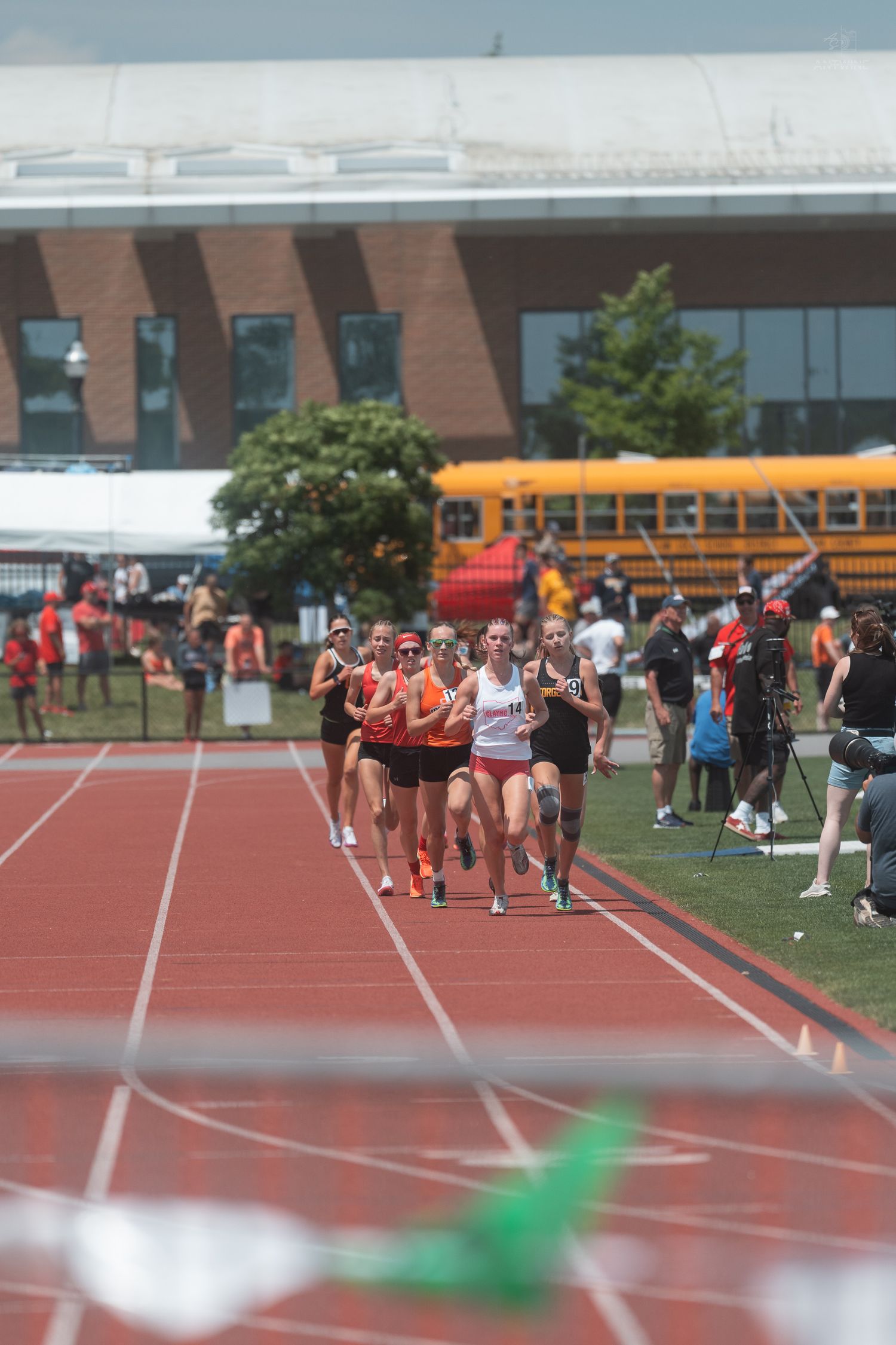 Female runners in orange and white uniforms compete in a track race past spectators at an outdoor stadium.