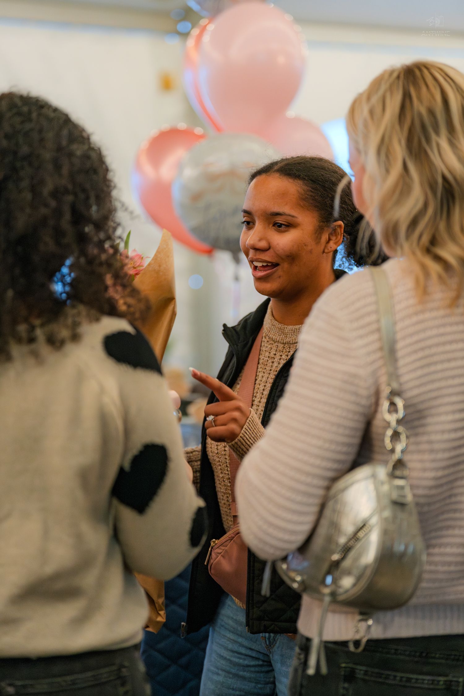 A smiling young woman in a pink sweater chats with friends near pink and silver balloons at a party.