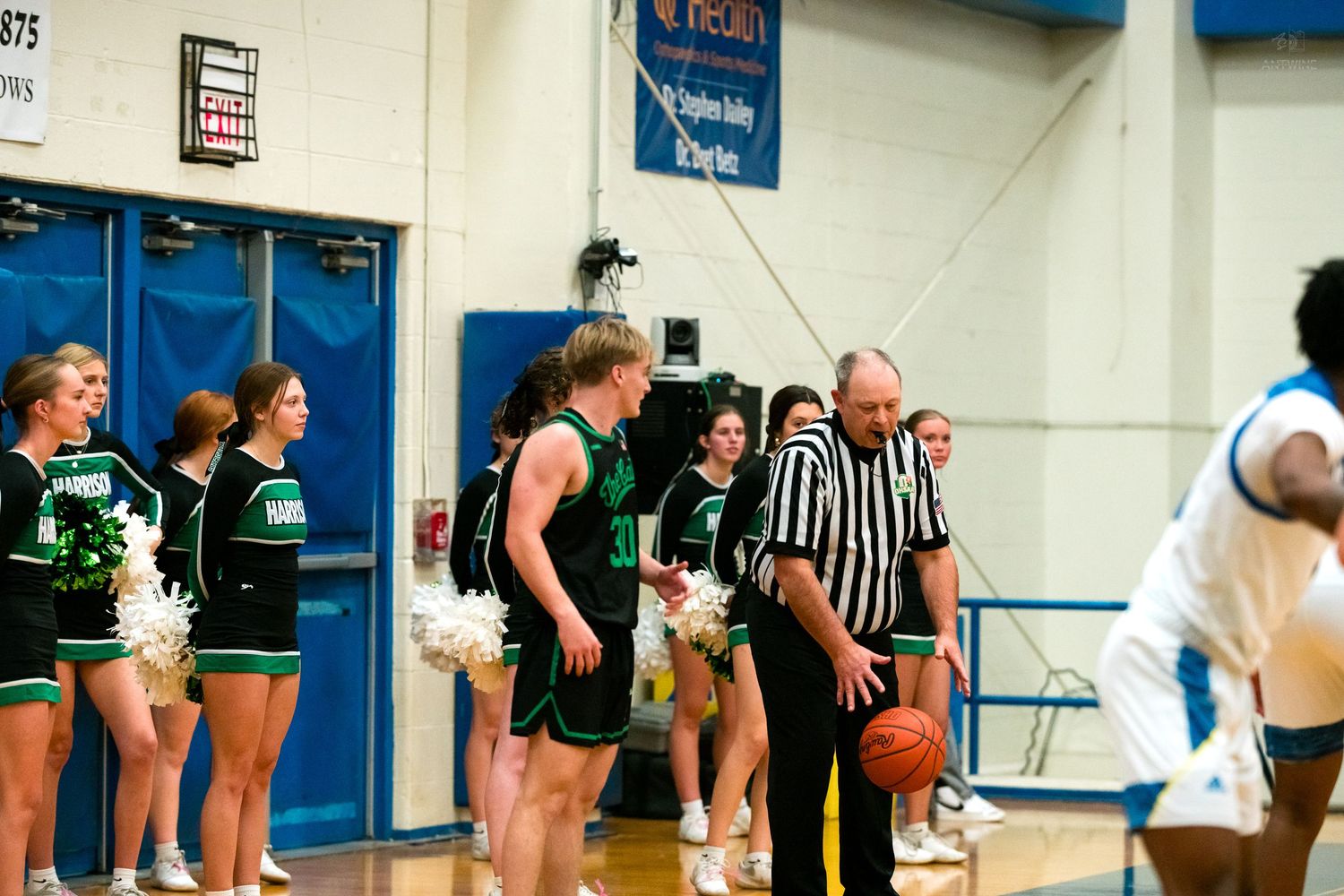 Green uniformed basketball players confer with referee near cheerleaders during indoor game.