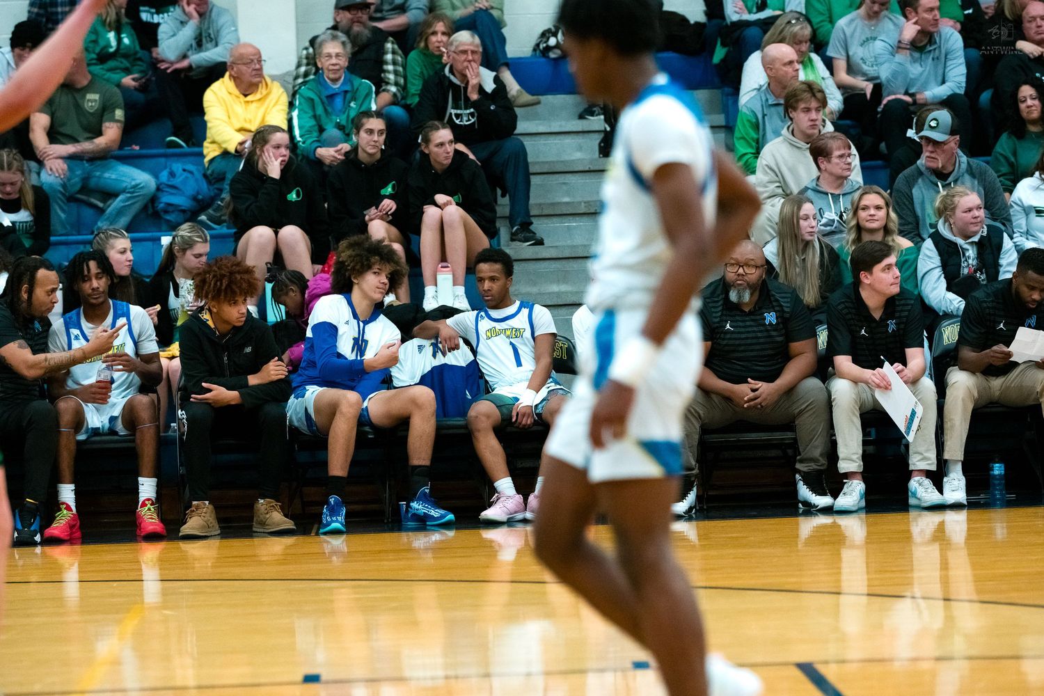 High school basketball players and coaches watch from the bench during an intense game against a green-uniformed team.