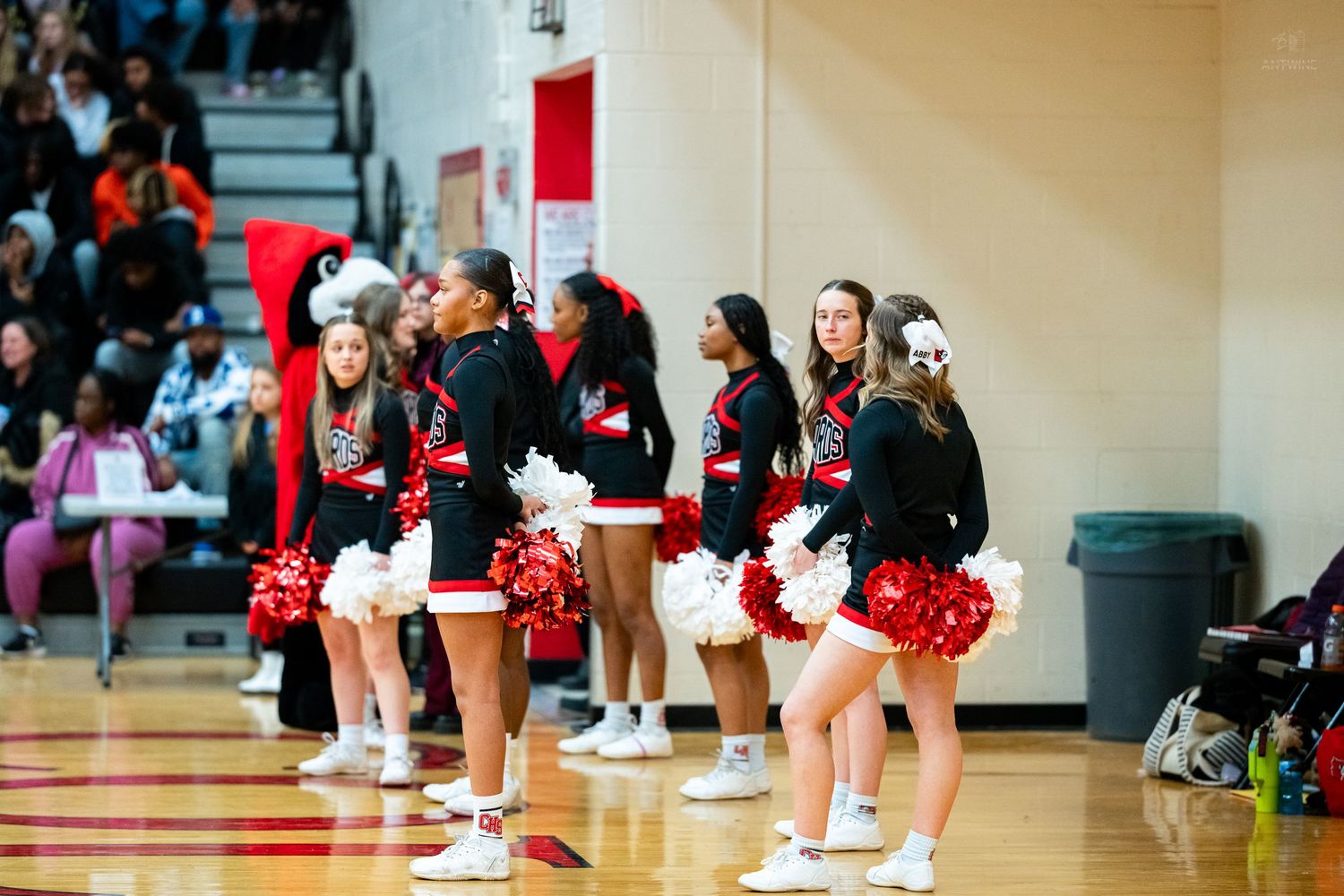 High school cheerleaders in black and red uniforms perform with pom-poms during indoor basketball game.