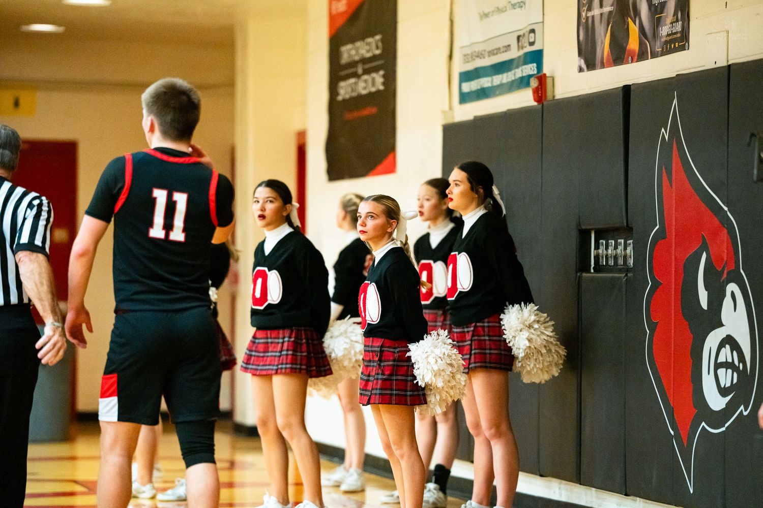 A basketball player in black jersey number 11 walks past cheerleaders in plaid skirts along the sideline.