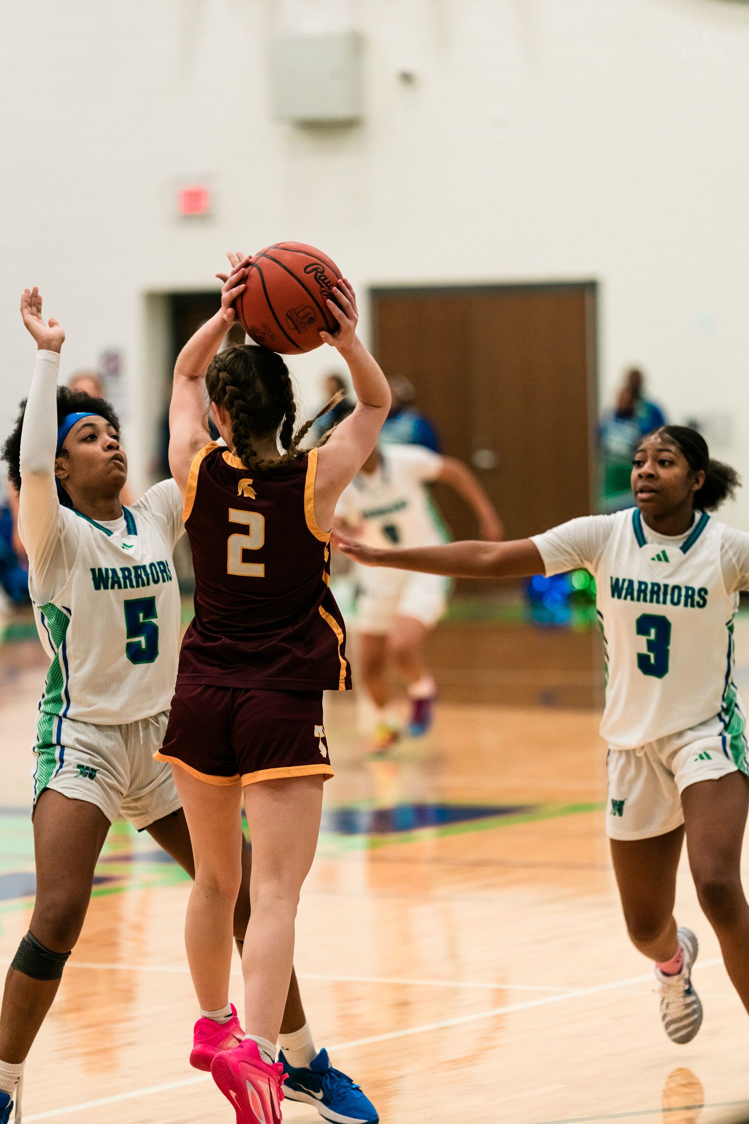 Two Warriors defenders in teal pressure a maroon-uniformed player number 2 attempting a shot in girls basketball.