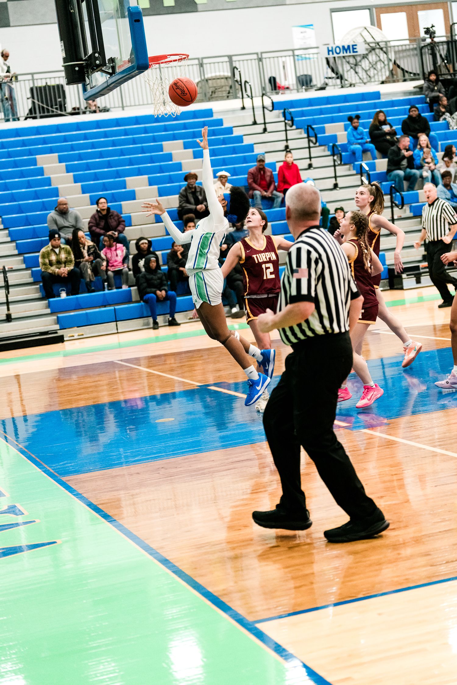 A player in a green uniform attempts a jump shot while a referee and opposing players watch during a girls basketball game.