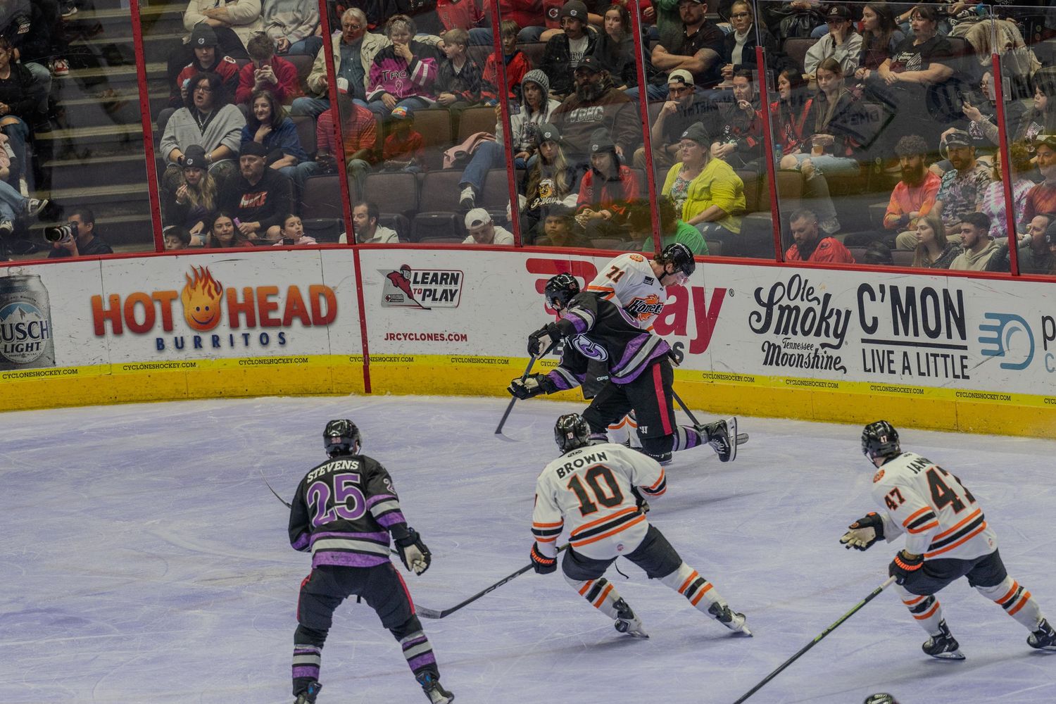 Ice hockey player leaps over opponent during intense gameplay with fans watching in arena.