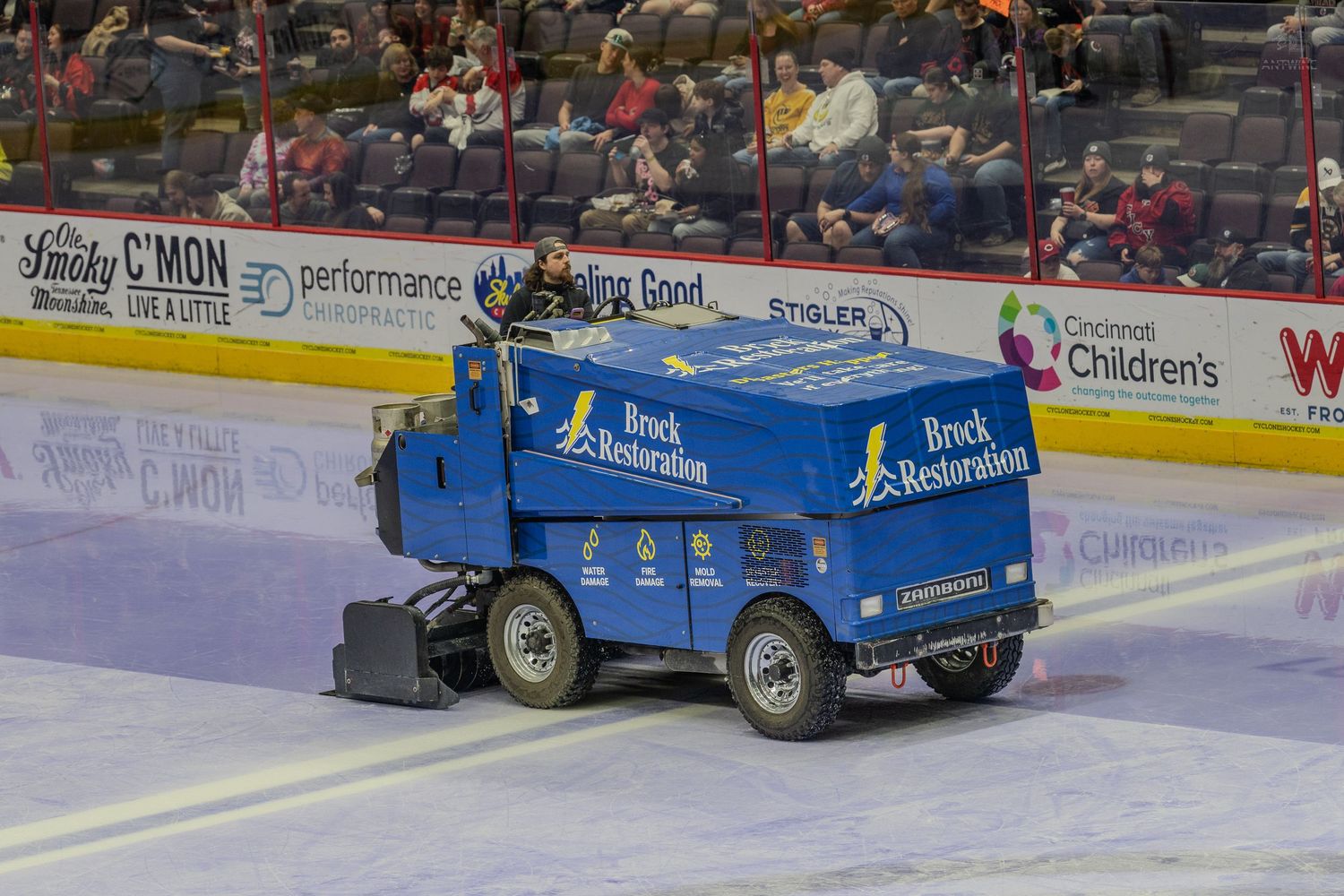 A blue Brock Restoration branded Zamboni resurfacing the ice at a hockey arena during an intermission.