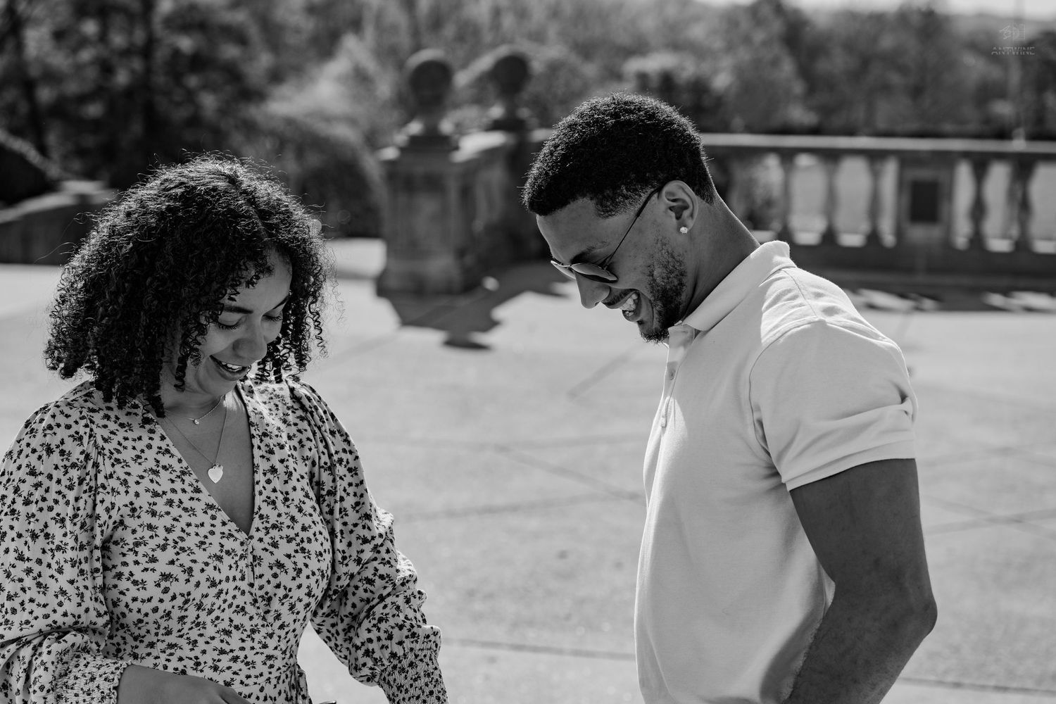 Black and white photo of a smiling couple looking down together on a sunlit terrace with stone balustrade.