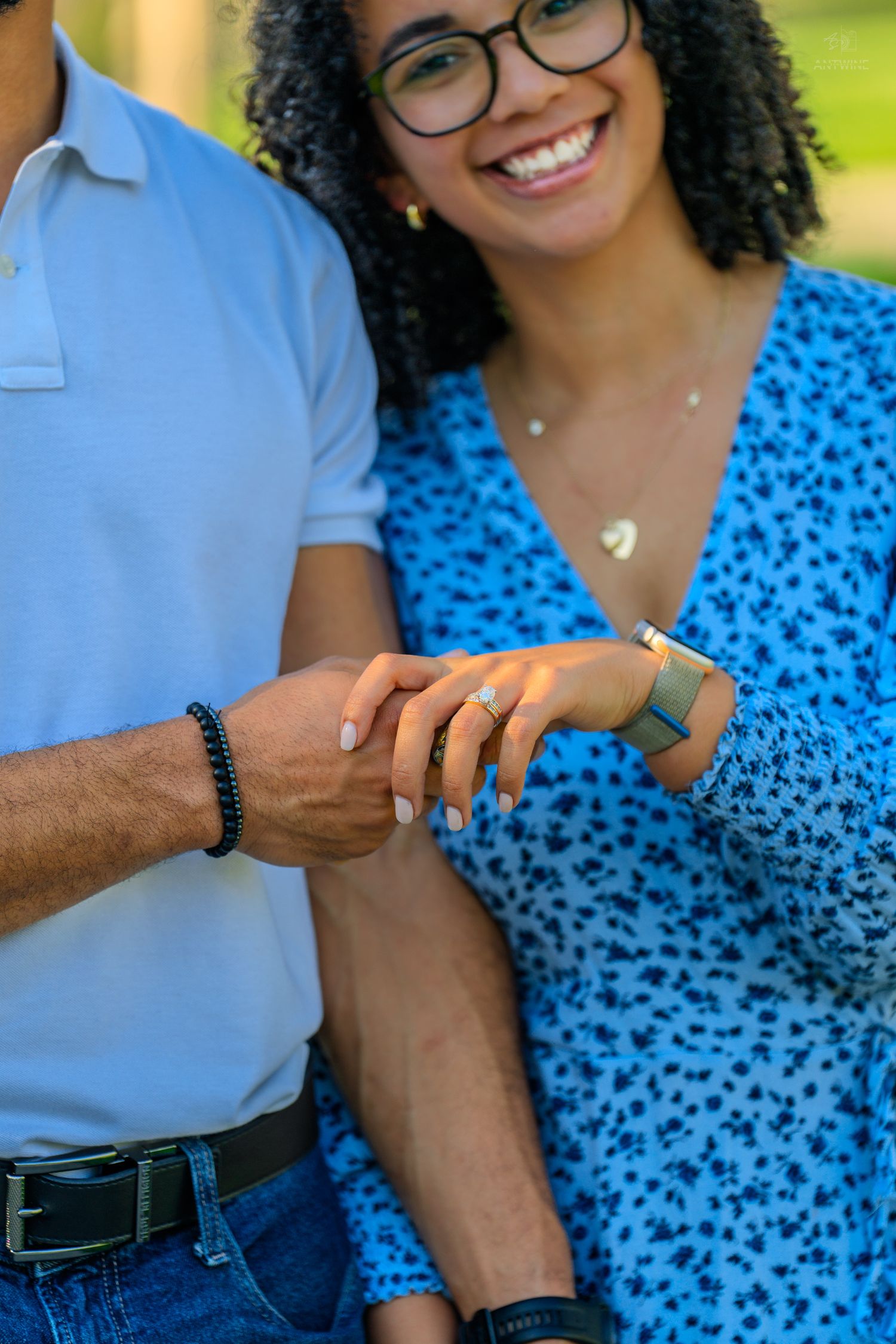 Close-up of a smiling woman in a blue dress showing off an engagement ring while holding her partners hand.