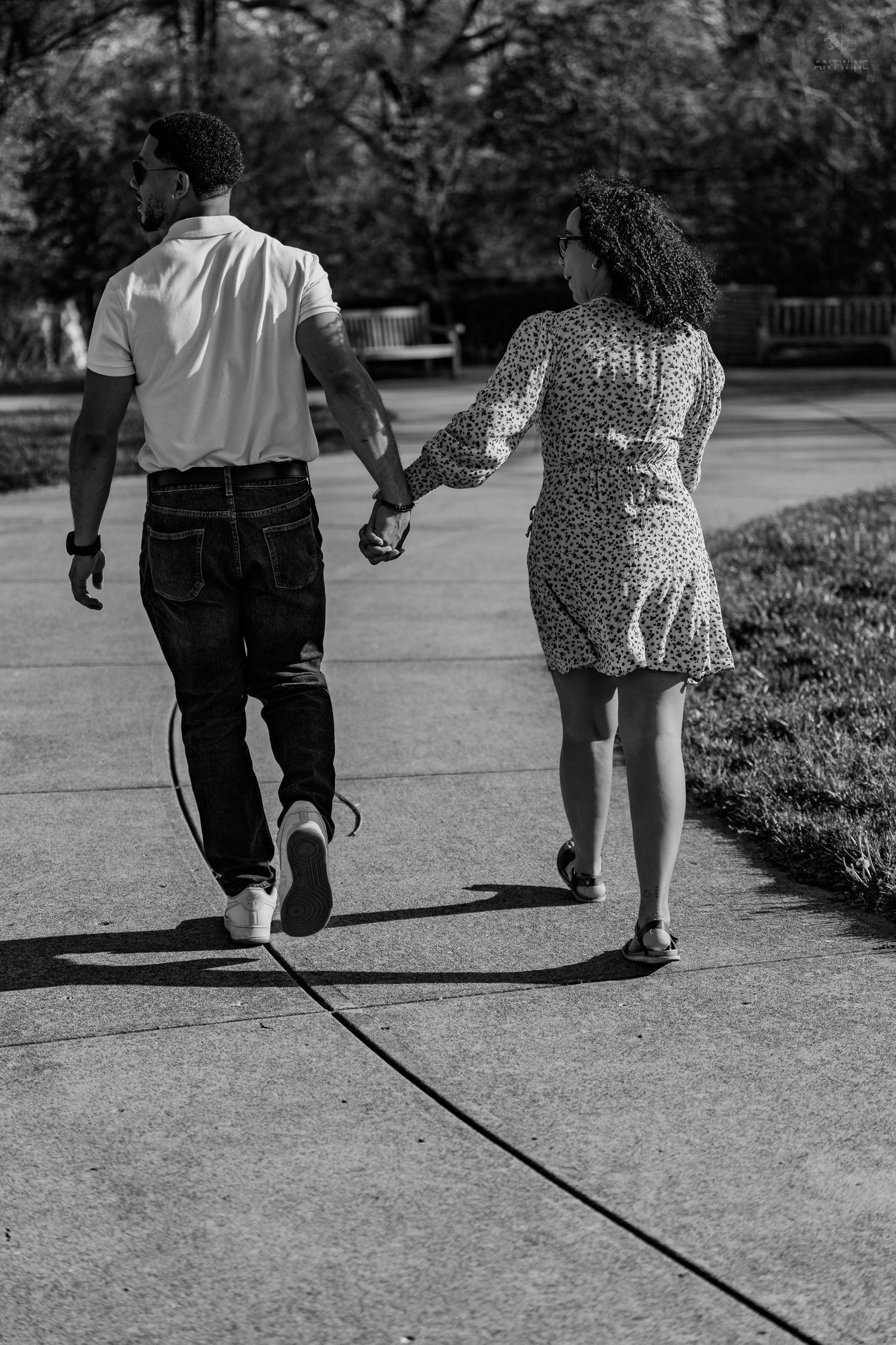 Black and white rear view of a couple holding hands walking along a park sidewalk.