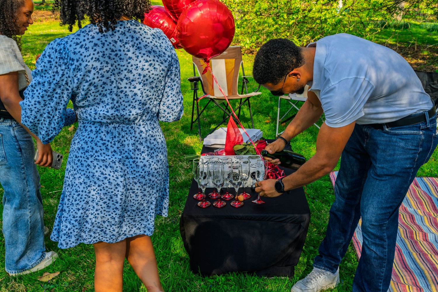 Man arranging champagne glasses on black table with red balloons at outdoor garden celebration.