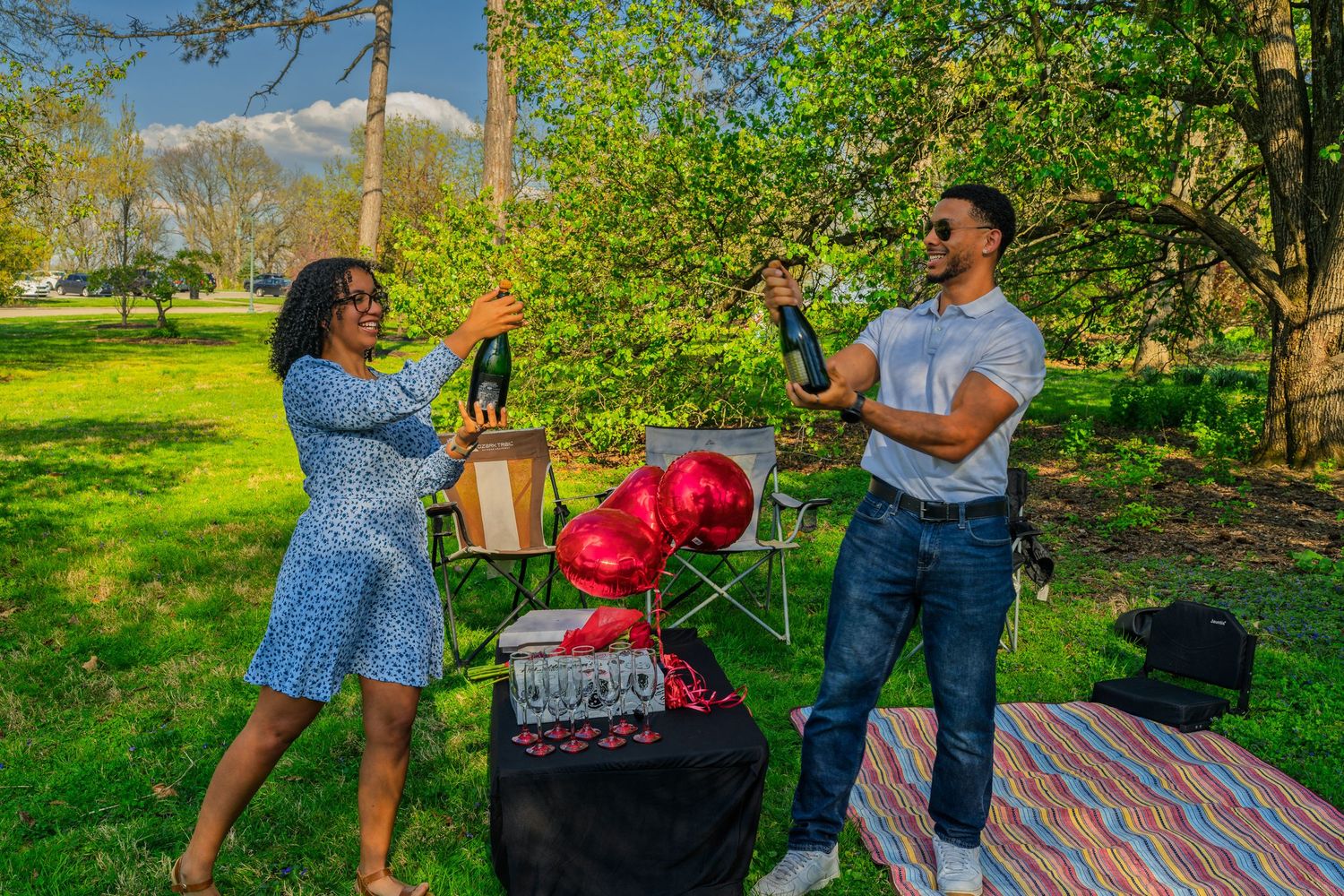 Couple celebrating outdoors holding champagne bottles with red heart balloons during a romantic park picnic.