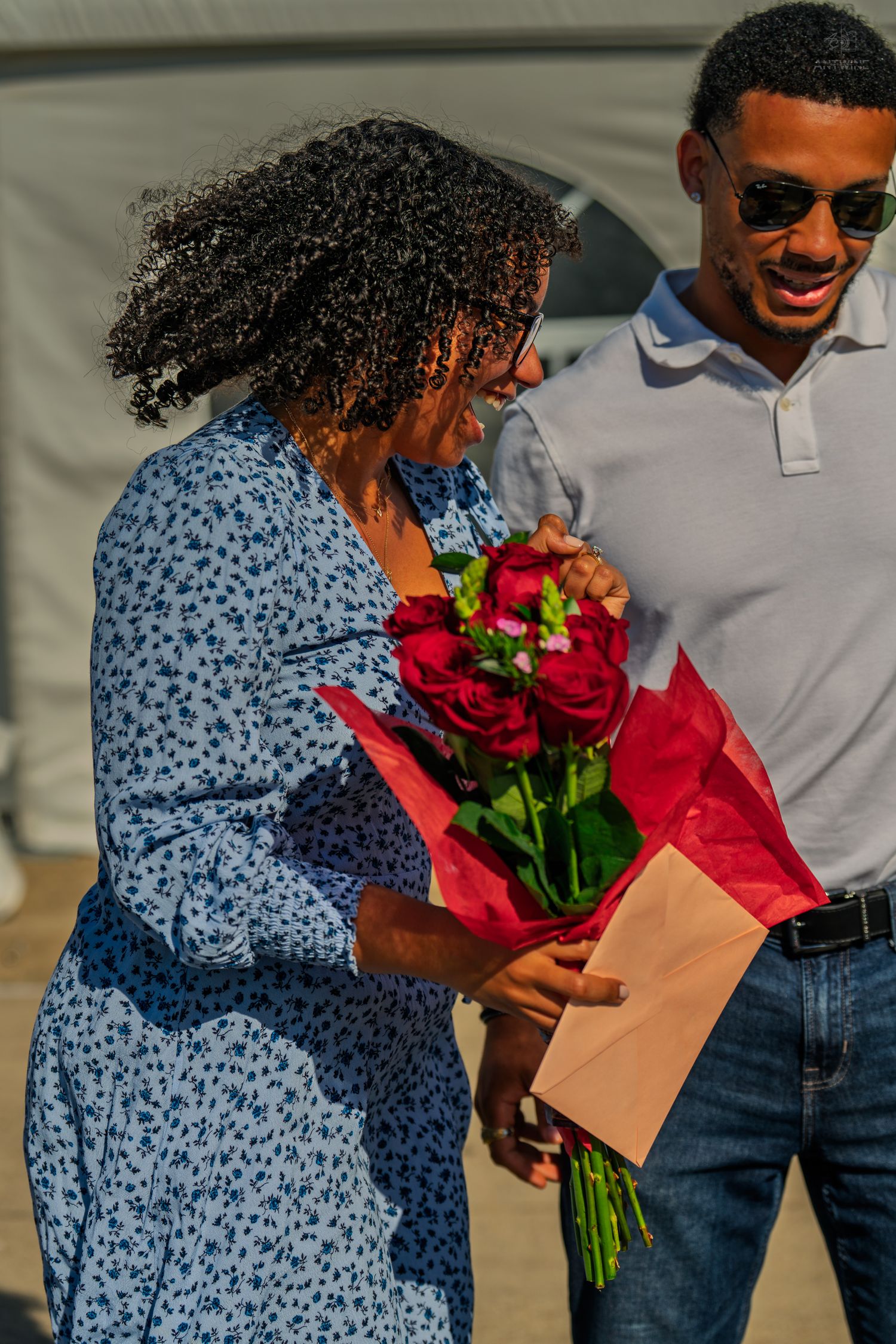 A man in sunglasses smiles as a woman in a blue dress admires a bouquet of red roses and an envelope.
