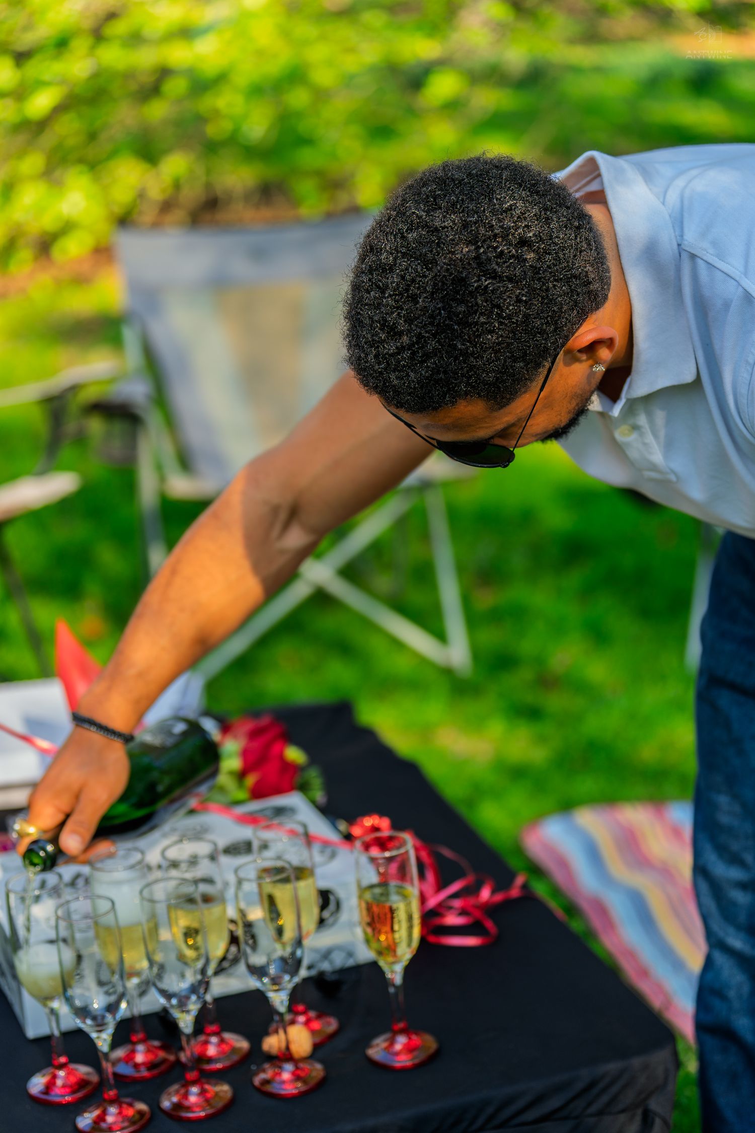 Man pouring champagne into flutes arranged on black table at outdoor garden celebration event.