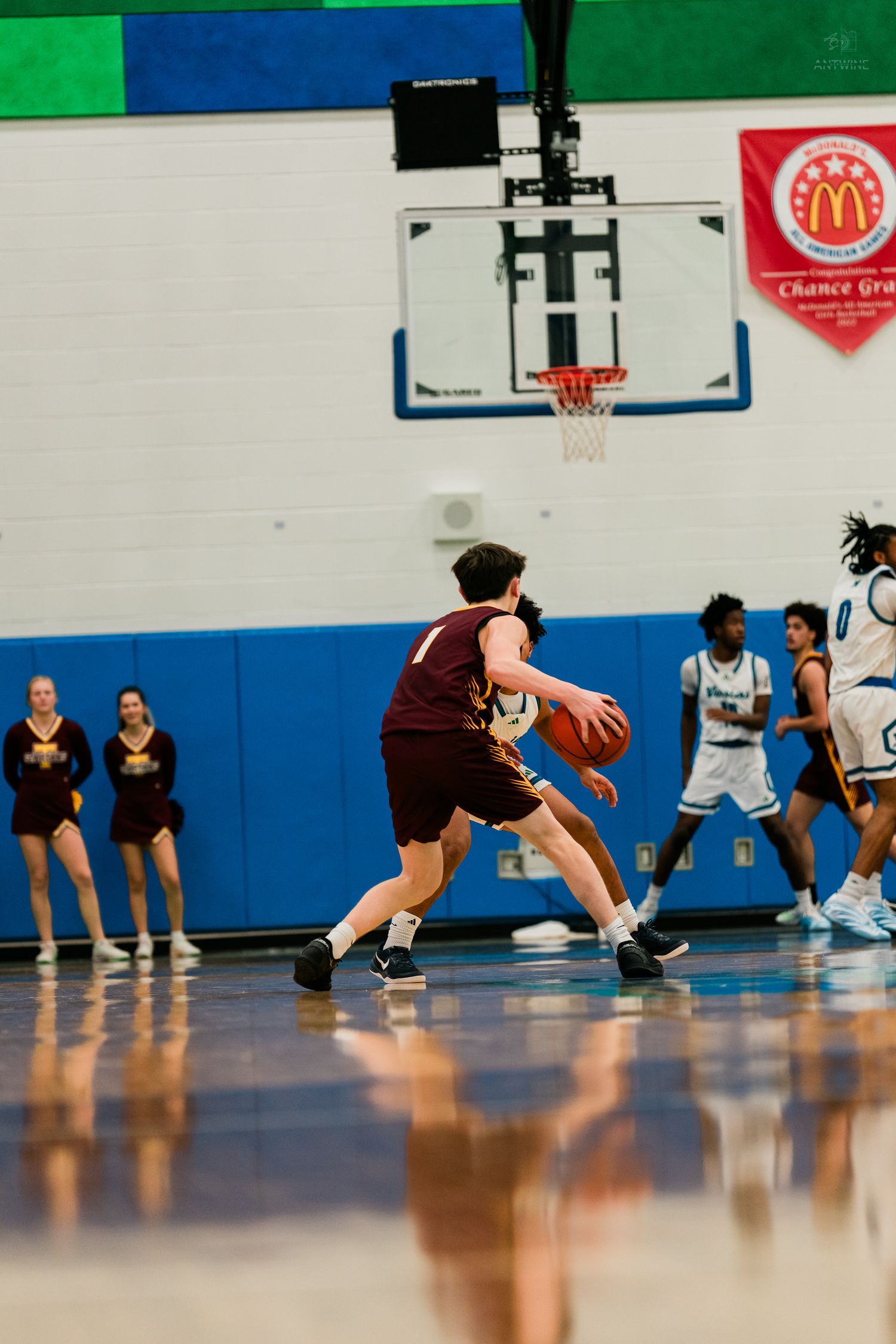 A maroon-uniformed player dribbles near the baseline while defenders in white close in during a basketball game.