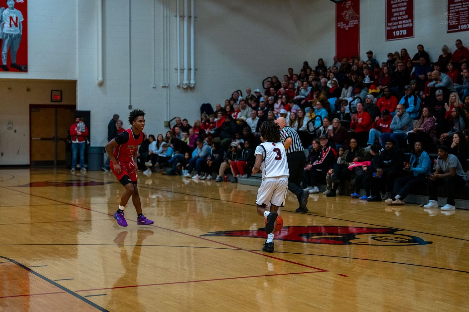 High school basketball player in white jersey dribbles ball while defender in red guards during game.