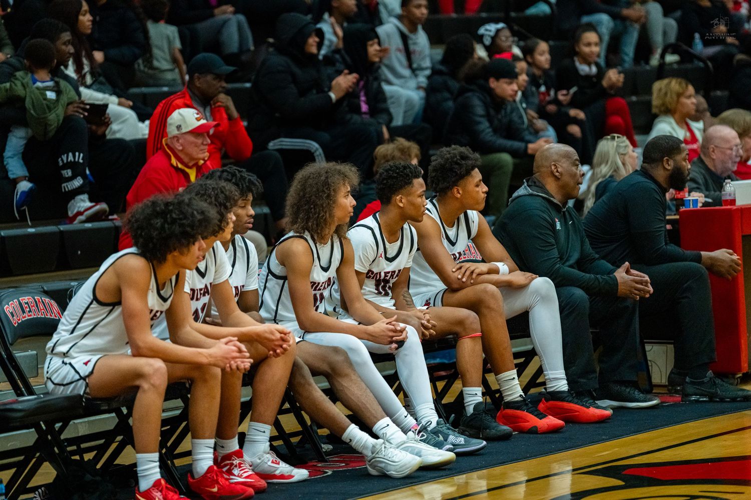 High school basketball players sit on the bench watching the game alongside coaches in a packed gym.