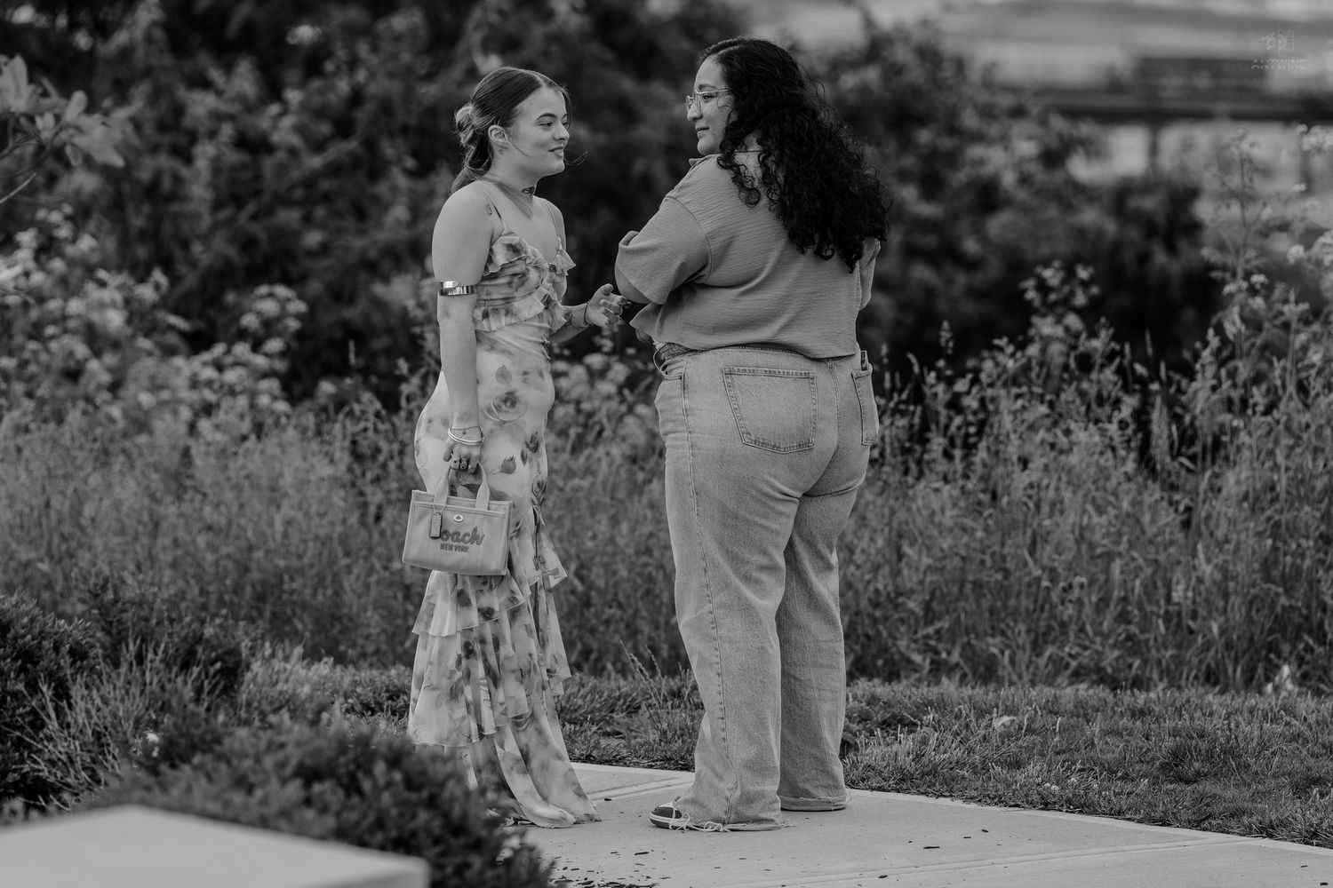 Black and white photo of two women conversing on an outdoor garden path surrounded by lush greenery.