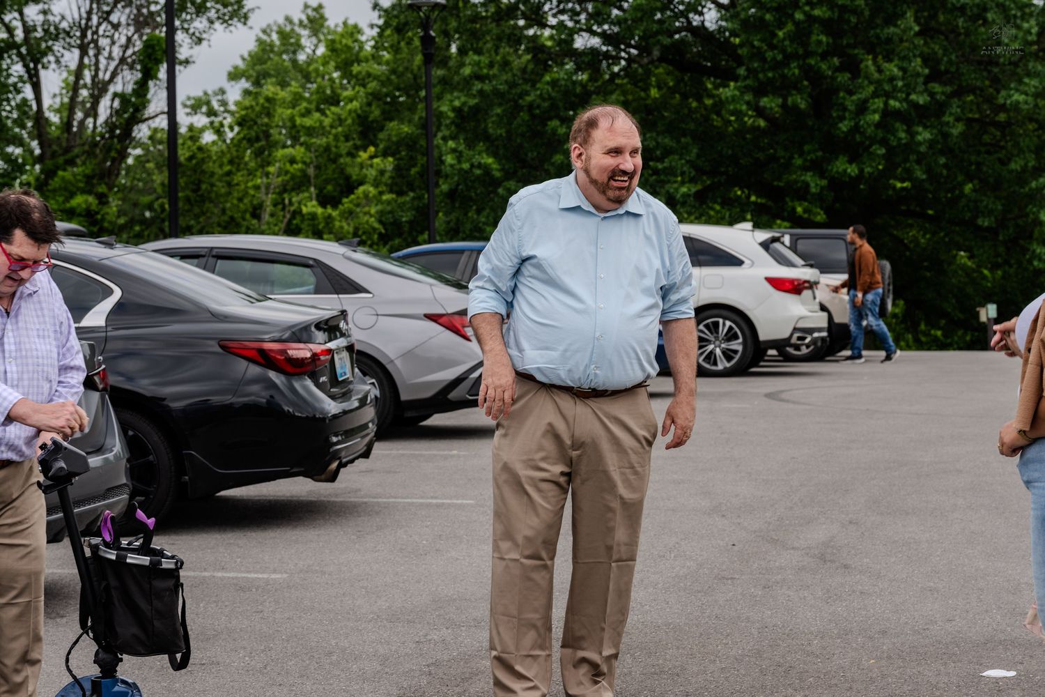 Man in blue shirt and khaki pants smiling in a parking lot surrounded by cars and trees.