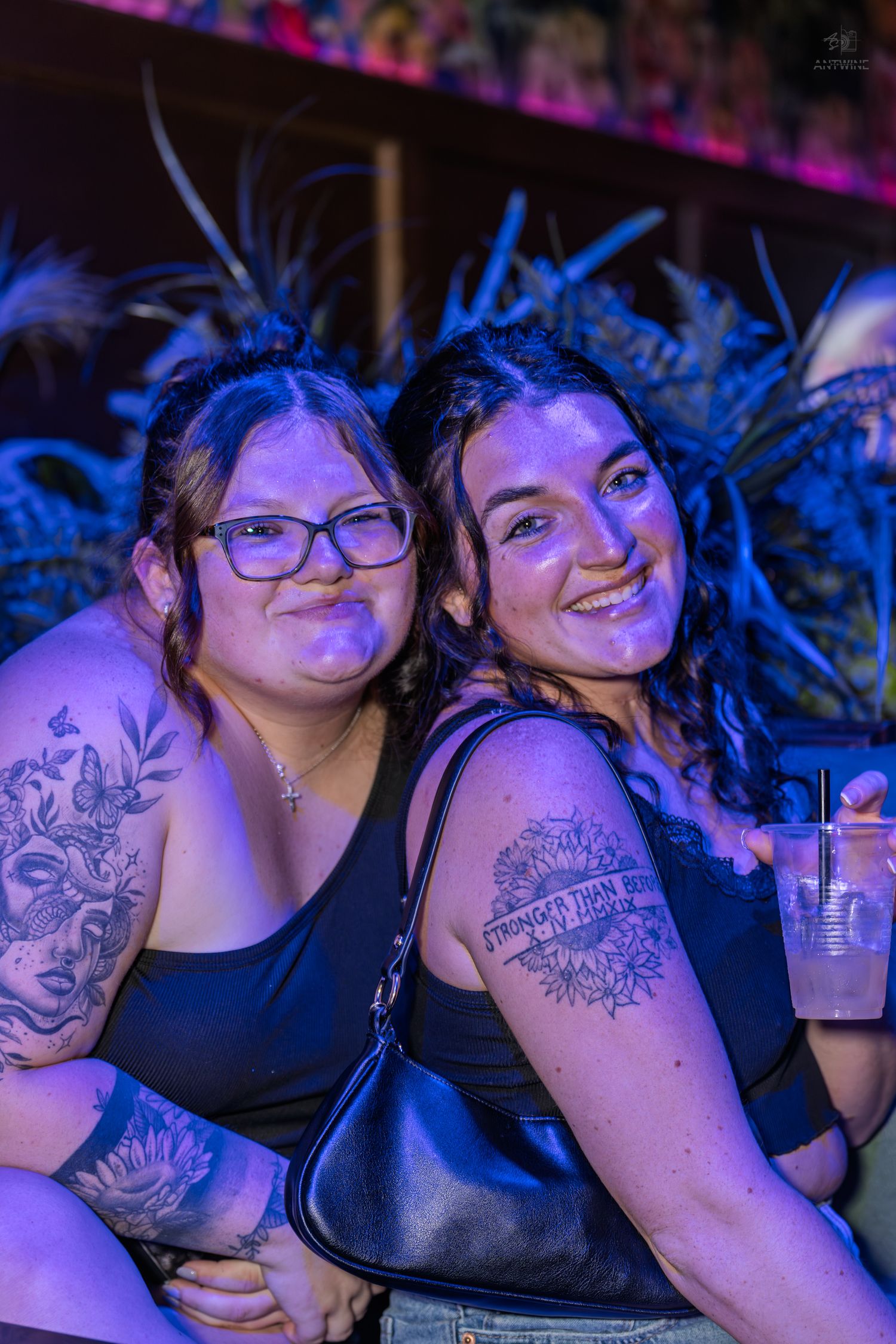 Two tattooed women in black tops smile together under purple nightclub lighting while holding a drink.
