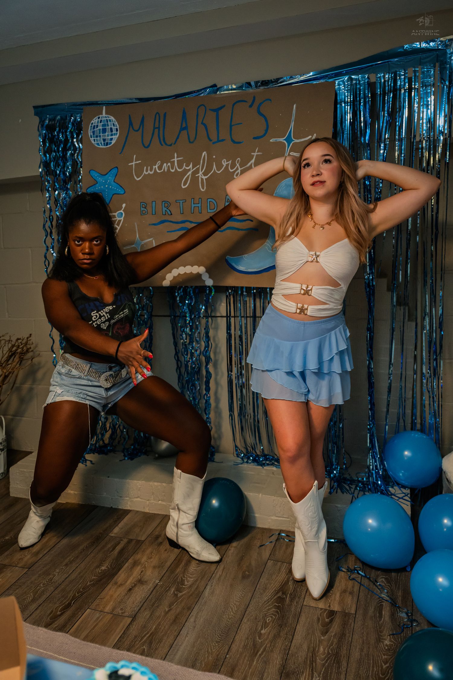 Two women strike poses at a blue-themed 21st birthday celebration with balloons and tinsel decor.