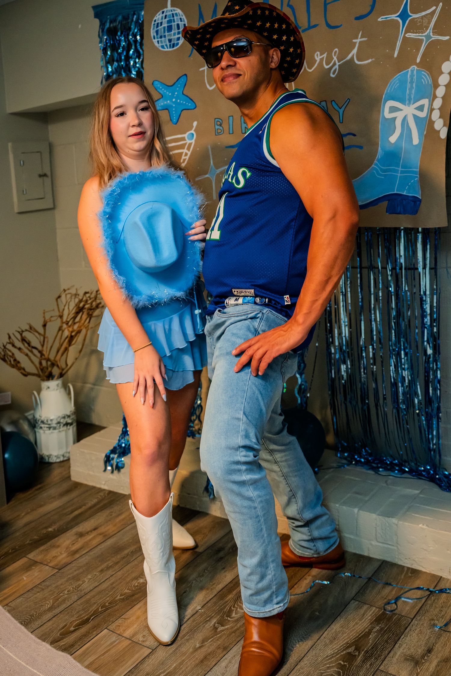 Young woman holds blue cowboy hat beside muscular man in Dallas jersey at birthday celebration.