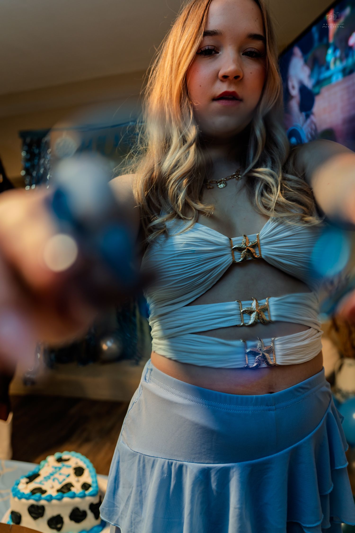 Young woman in blue crop top and skirt pointing at camera at a birthday party.