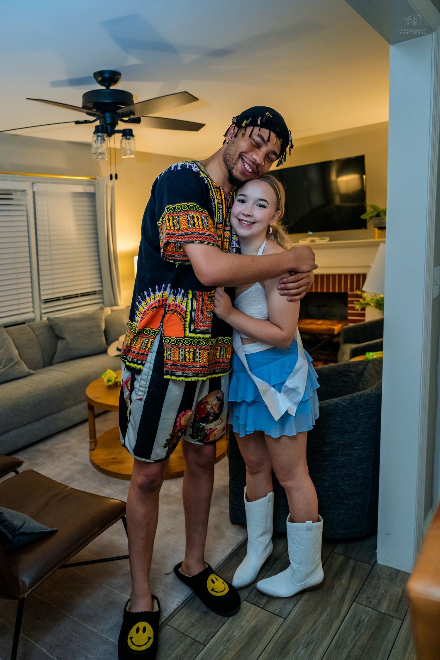Young man in dashiki and woman in blue skirt and white boots share a warm hug indoors.