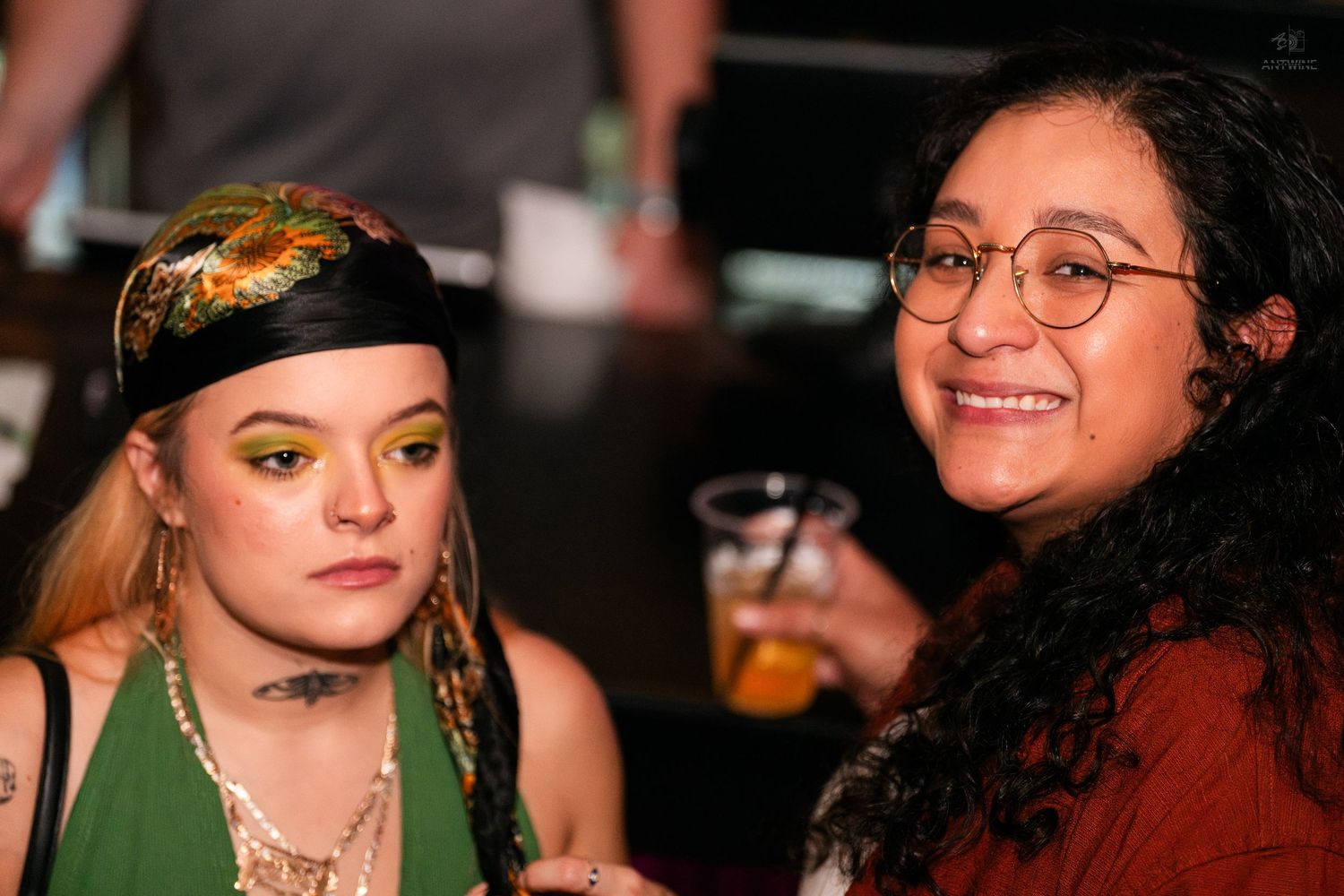 Two women smiling together at a bar, one wearing a headband and green top.