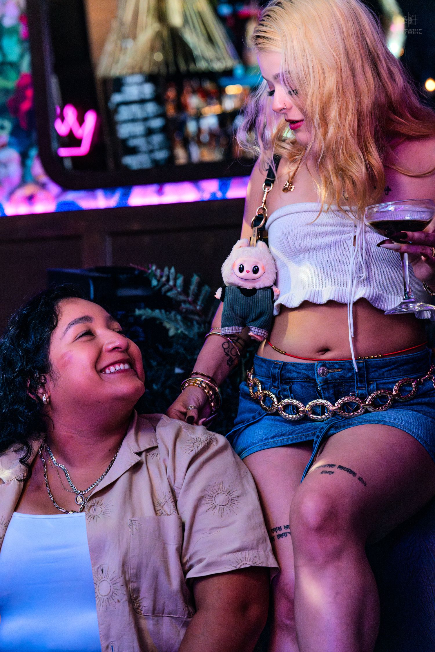 Two women laughing together at a neon-lit bar, blonde holding wine glass wearing white crop top and denim shorts.