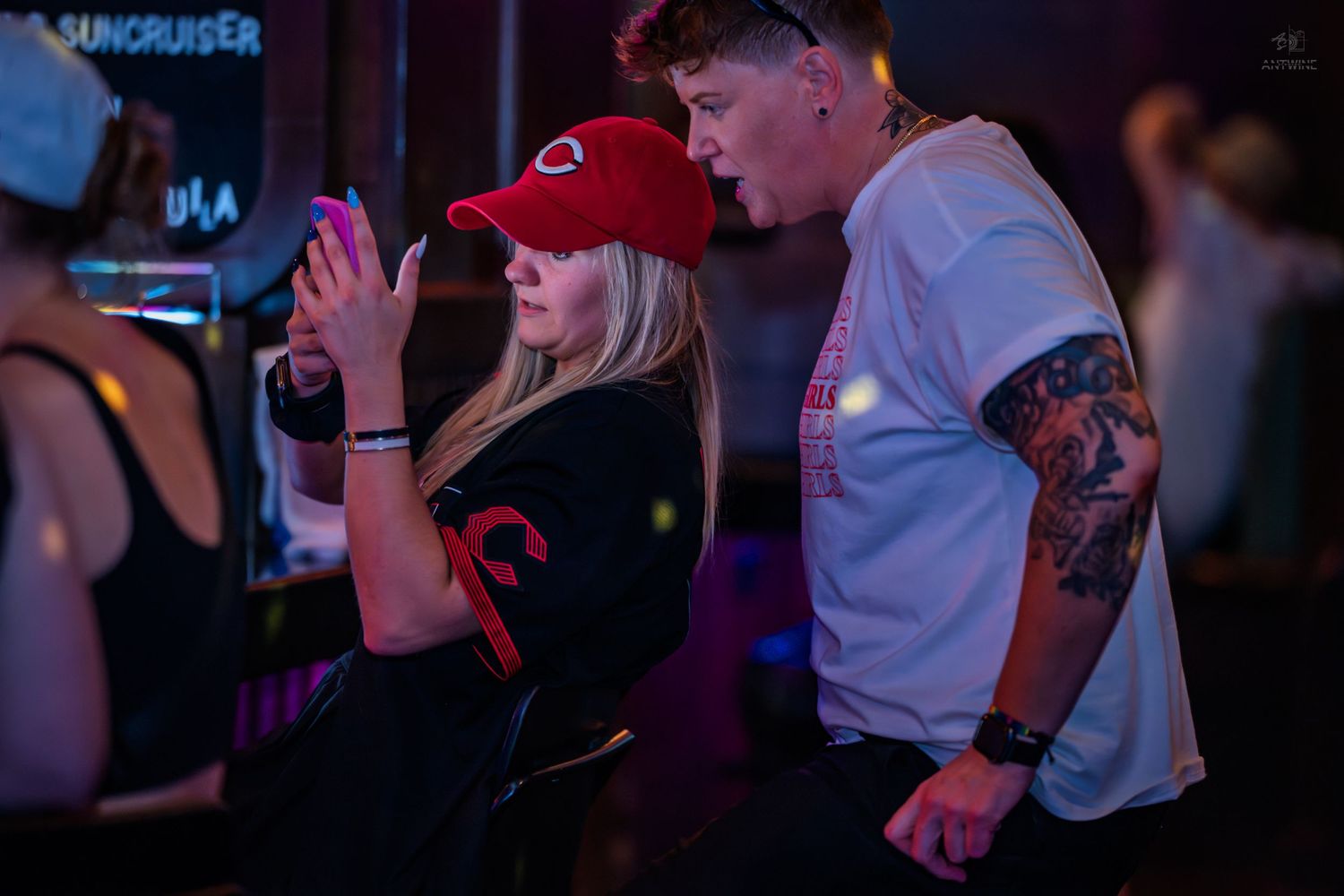 Blonde woman in red cap using phone at a bar with tattooed man leaning in.
