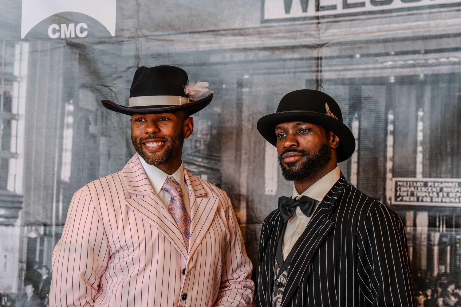 Two well-dressed men in vintage pinstripe suits and fedora hats posing together at a formal event.
