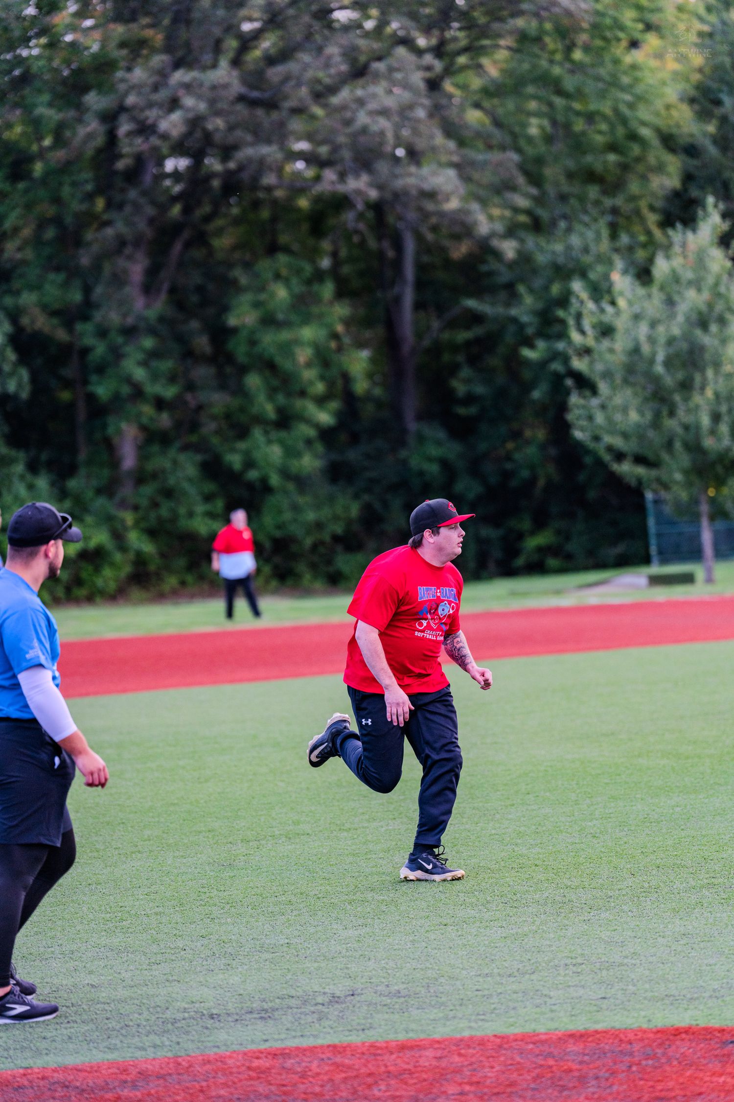 Player in red shirt running bases during softball game with umpire watching on green turf field.